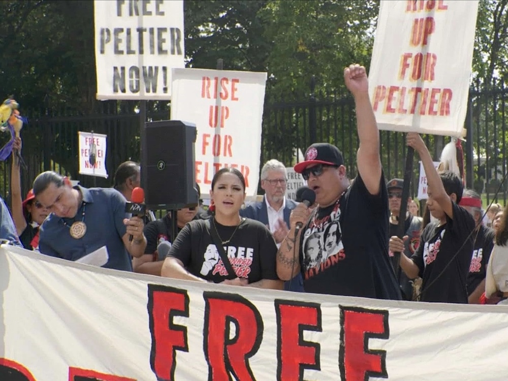 The front of a protest line featuring Native American activists features a man and a woman in black graphic T-shirts behind a large white banner with the words &lsquo;Free Leonard Peltier.&rsquo; Others are holding protest signs behind them that say &lsquo;Free Peltier Now!&rsquo; and &lsquo;Rise Up for Peltier.&rsquo;