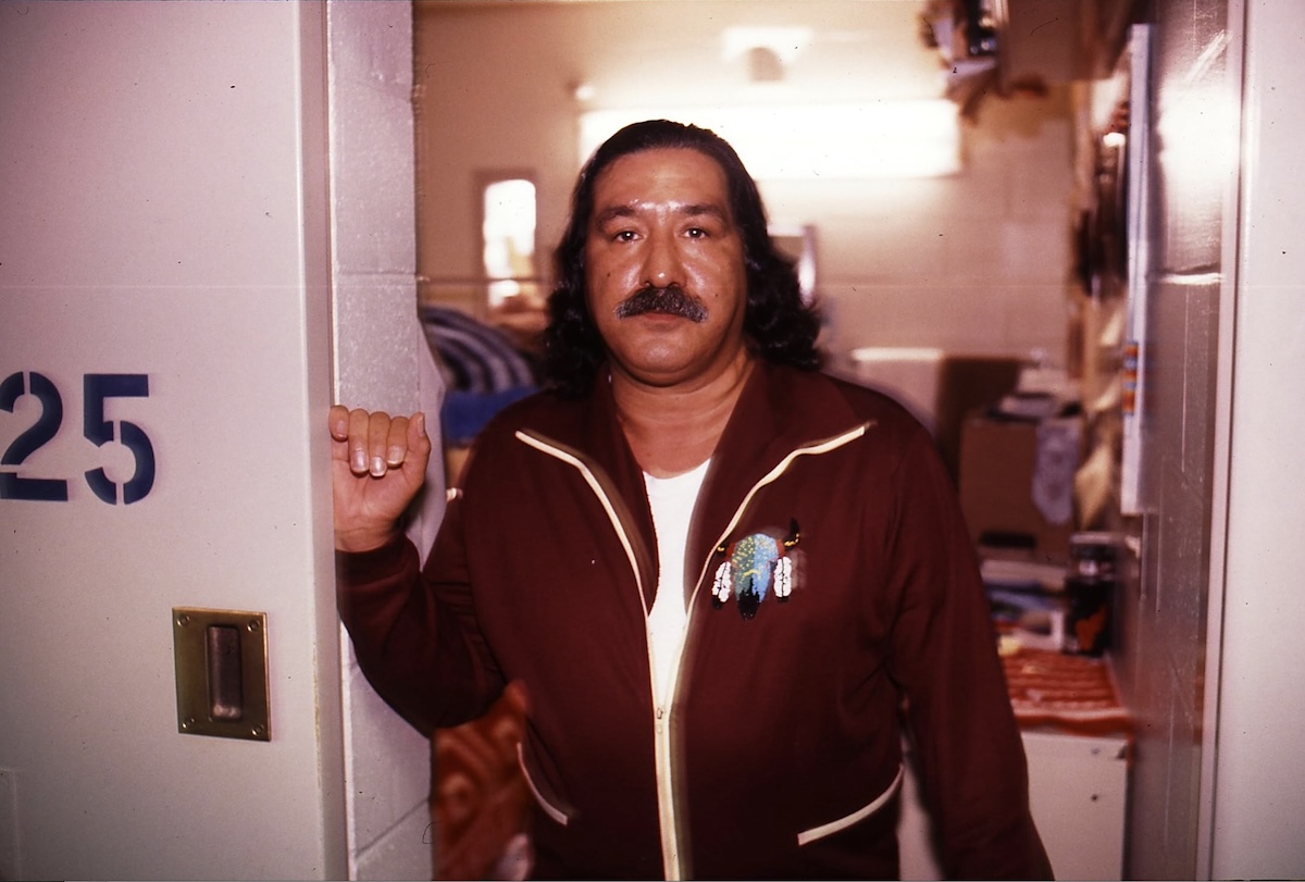 Leonard Peltier has dark, wavy, shoulder-length hair, a moustache and a medium skin tone. He is wearing a burgundy tracksuit over a white T-shirt. An embroidered image of a horned animal in a Native American style sits on the left side of his chest. He stands at a doorway of a room in an institution with white concrete walls. 
