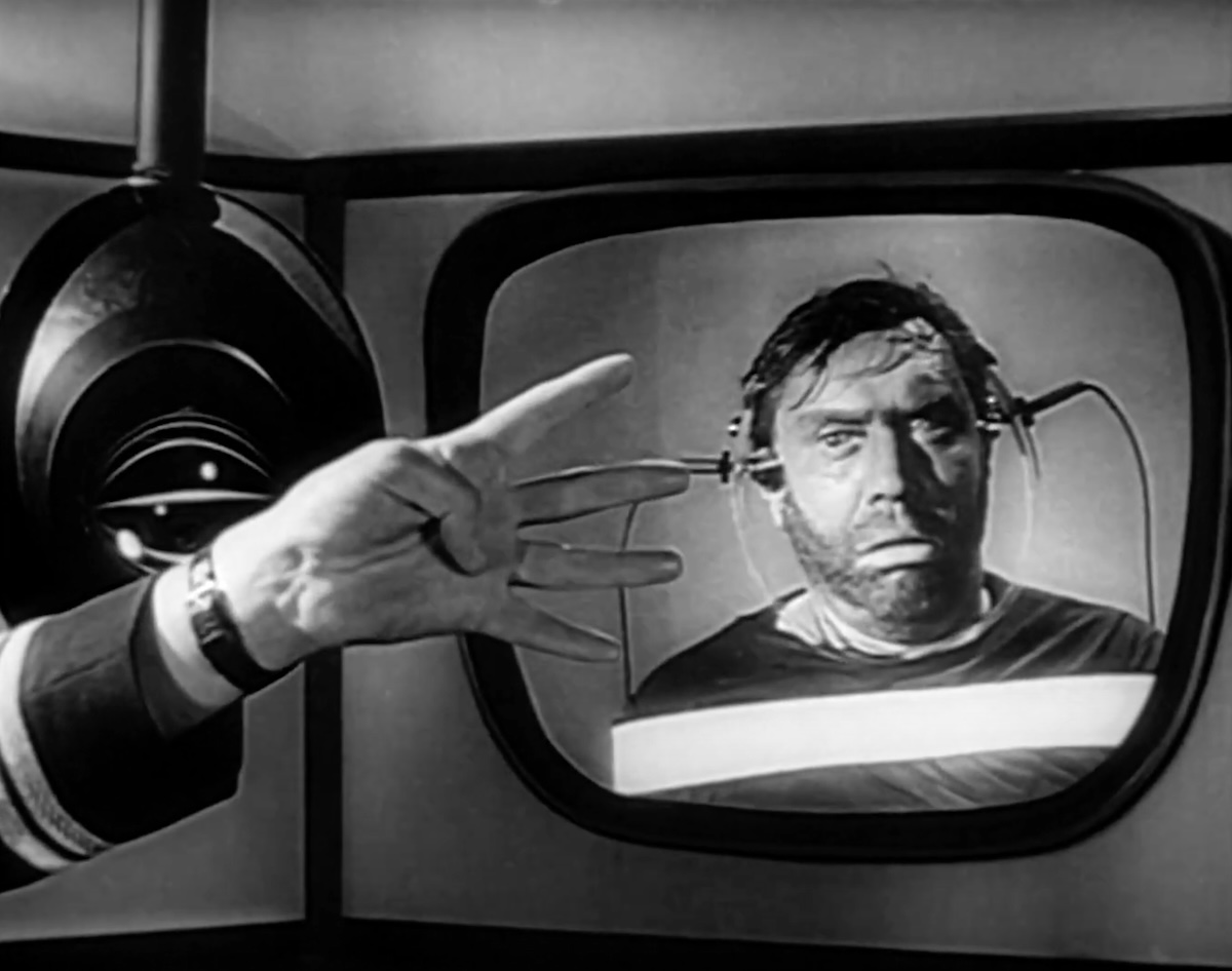 A black and white film still depicts a hand holding up four fingers and a man on a projector screen looking on, wearing headphones.