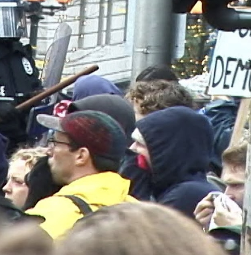 People in casual streetwear are assembled in a crowd, protesting, on a grey day in down Seattle. Some are holding protest signs. They are facing a line of police officers in dark riot gear.