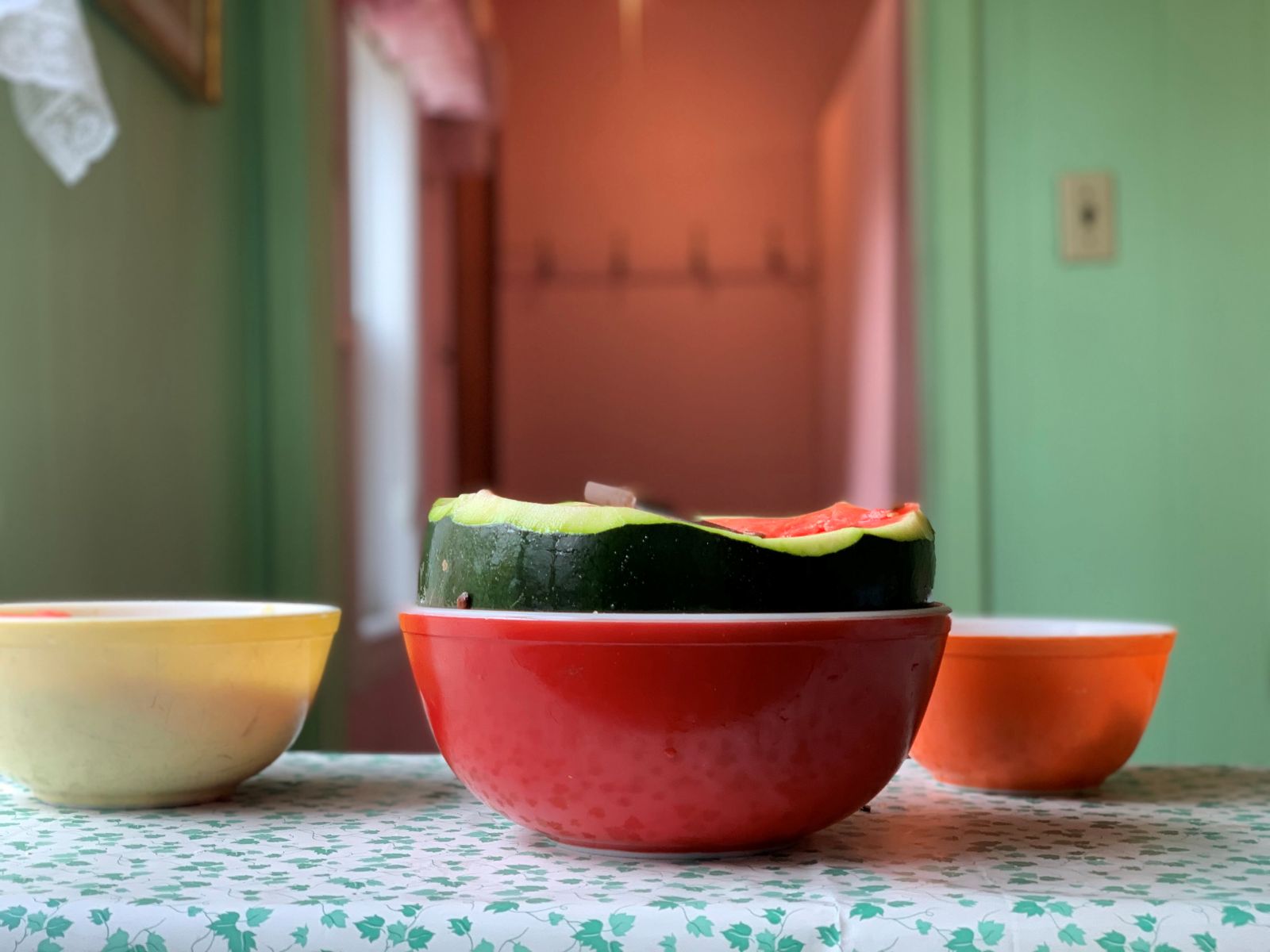 A half watermelon sits, skin side down, in a red bowl on a white and green leaf-patterned tablecloth next to a yellow and orange bowl in a kitchen.