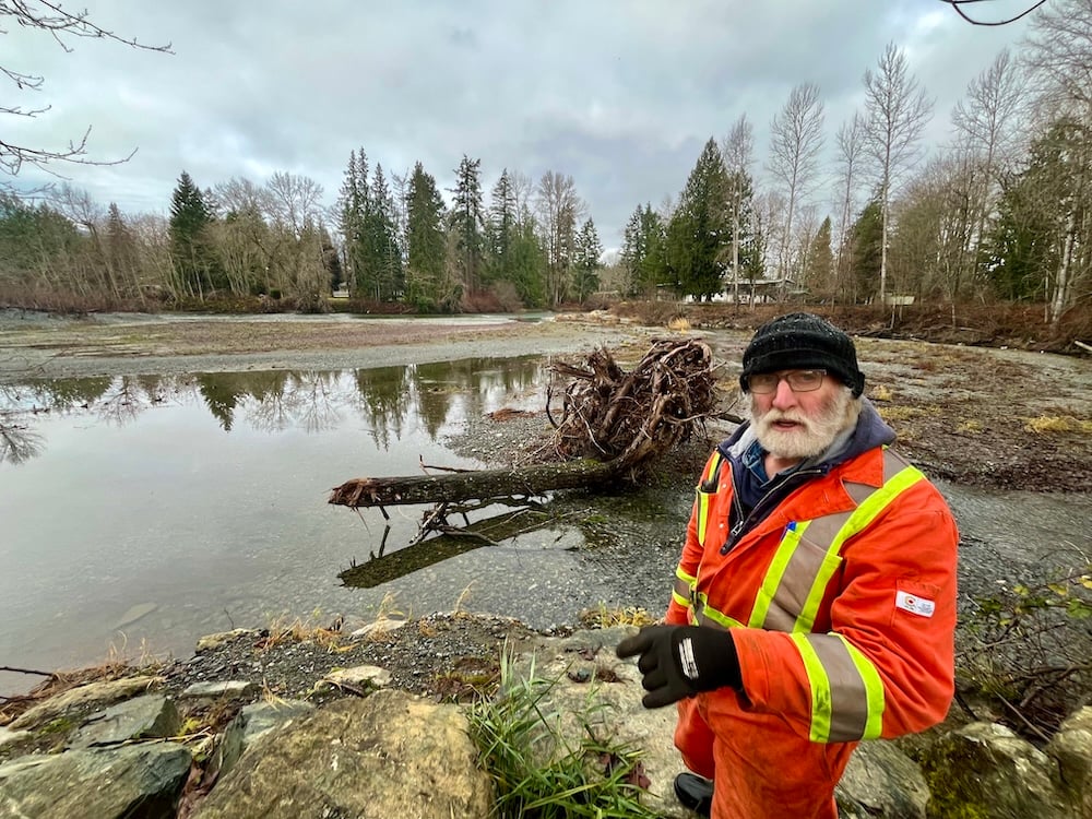 Colin James has a grey beard and is wearing a black toque and orange high-visibility coveralls. He is standing in front of a placid river, but a large uprooted tree suggests the area was recently flooded.