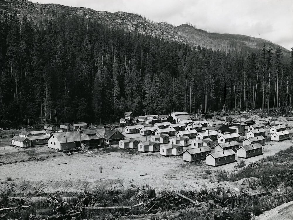 A black and white photo shows a worker camp featuring more than a dozen small modular homes along with several larger buildings.