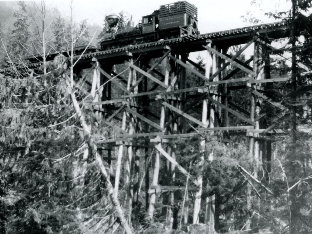 A black-and-white image of a steam train crossing a high wooden train trestle above the forest floor.