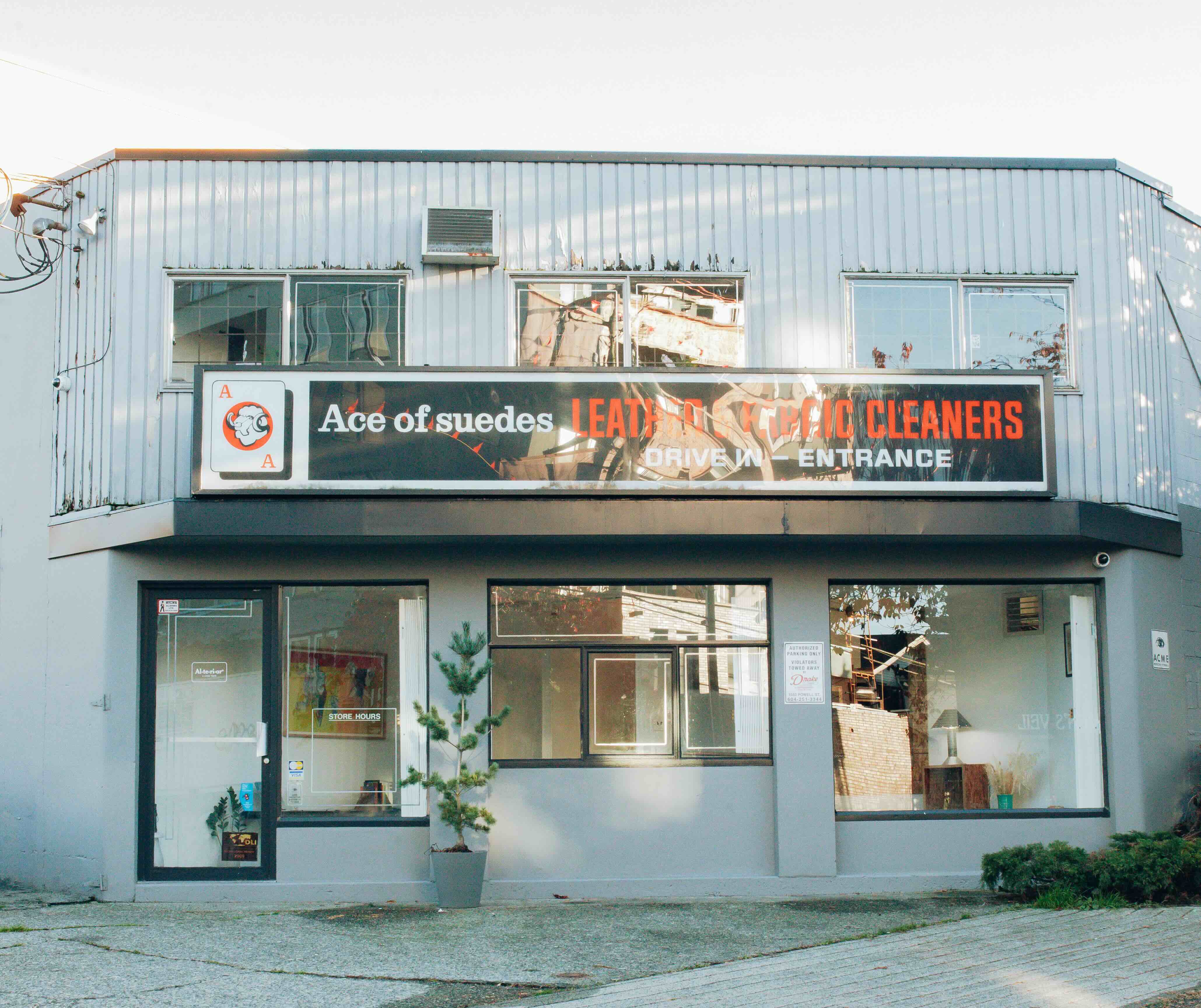 The exterior of a no-frills two-storey building painted light grey features an awning that reads &ldquo;Ace of Suedes Leather and Fabric cleaners&rdquo; in white and red text against a black background.