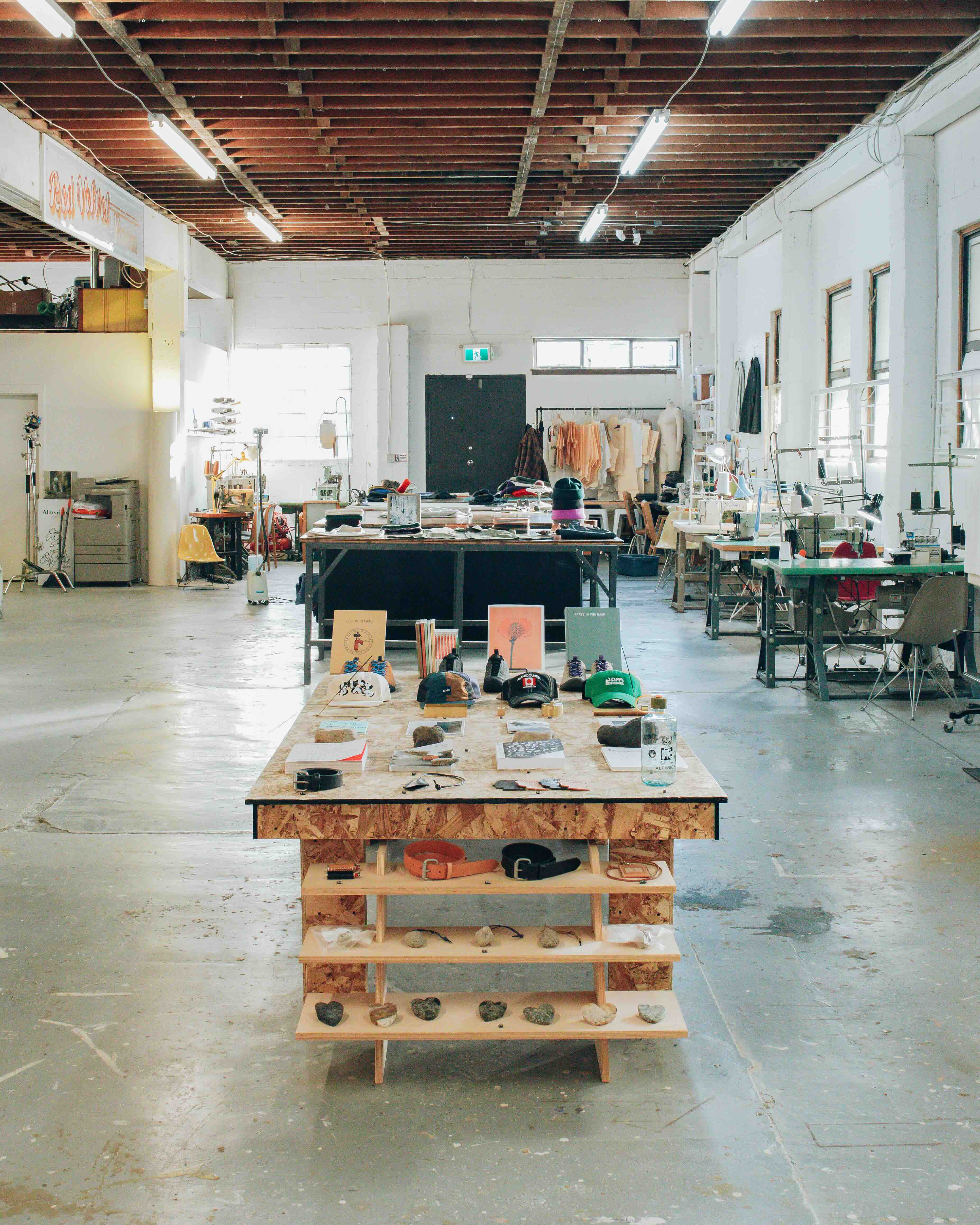 The interior of a retail and artisan space features hats, belts and other small retail items displayed on a wooden table in the middle of the frame. There are industrial grey floors and a row of sewing tables to the right in the background.