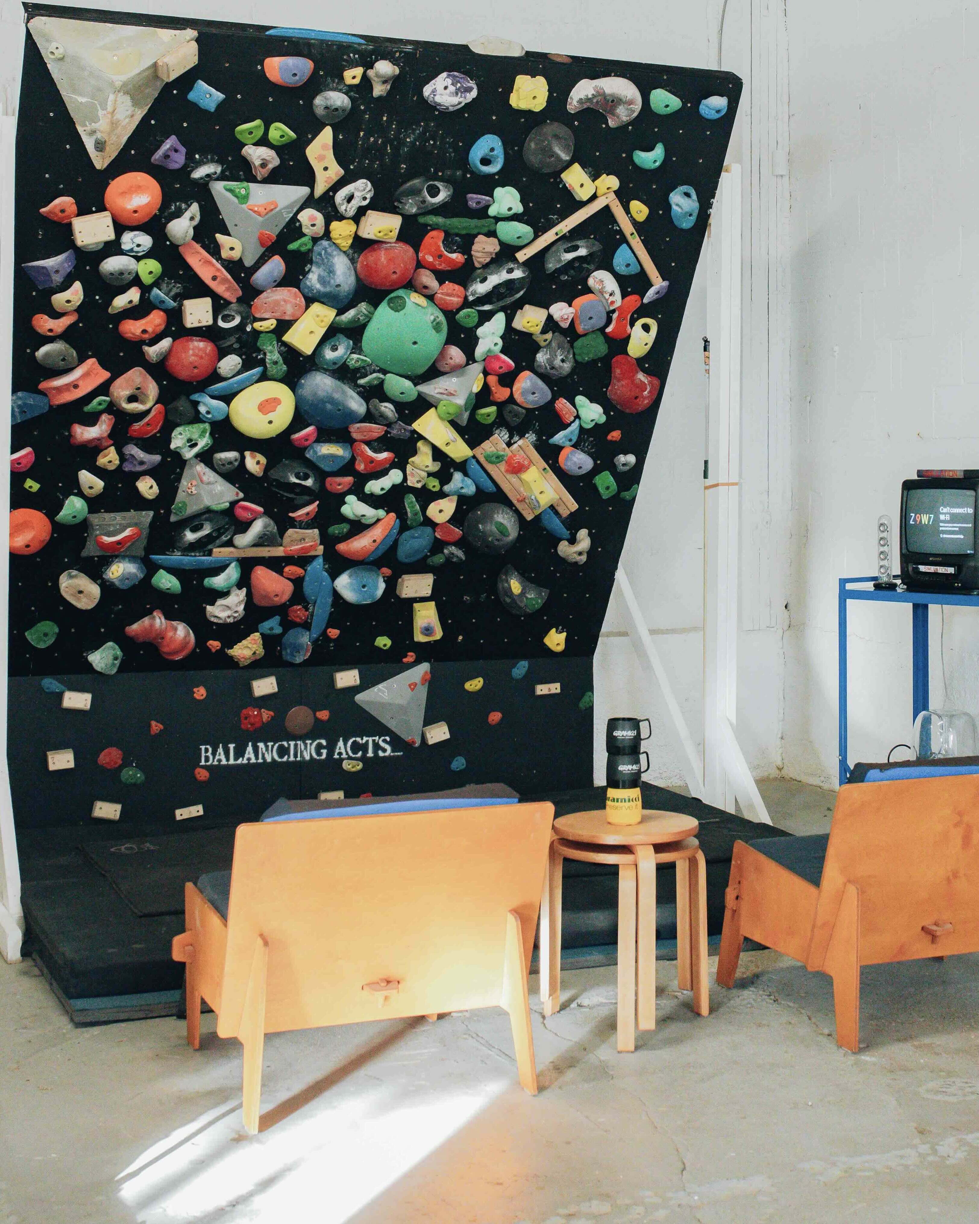 A black climbing wall bears a colourful assortment of indoor climbing rocks. In front of the wall are two low lounge chairs in a bright indoor space.
