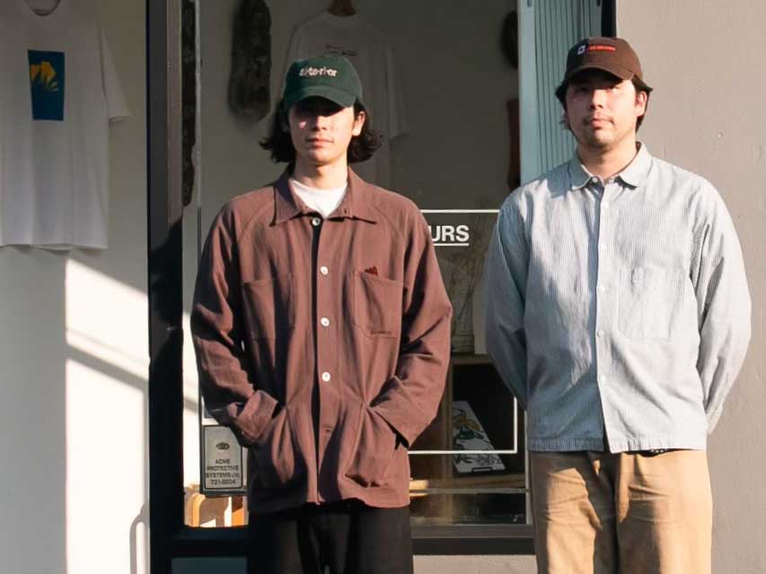 Dallas and Cole Chreptyk stand outside a storefront on a street. Dallas, left, is wearing a green ball cap and an oversized brown button-down shirt. Cole, right, is wearing a brown ball cap and a light blue button-down shirt. They are standing in the sun.