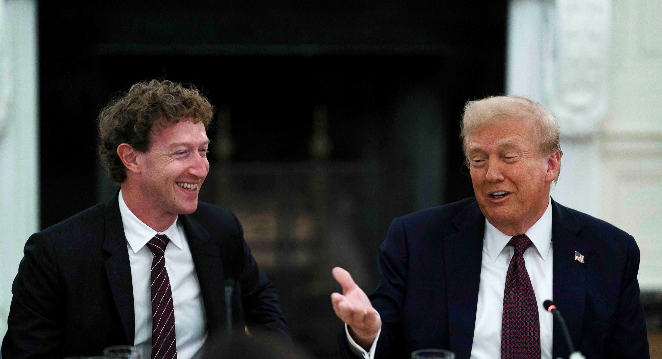 Two men in dark suits, white shirts and burgundy ties are seated at a dining table featuring light yellow floral centrepieces. Mark Zuckerberg, left, is a man in his 40s with curly brown hair. Donald Trump, right, is a man in his 70s with wavy dyed blond hair. They are speaking together, smiling and laughing.