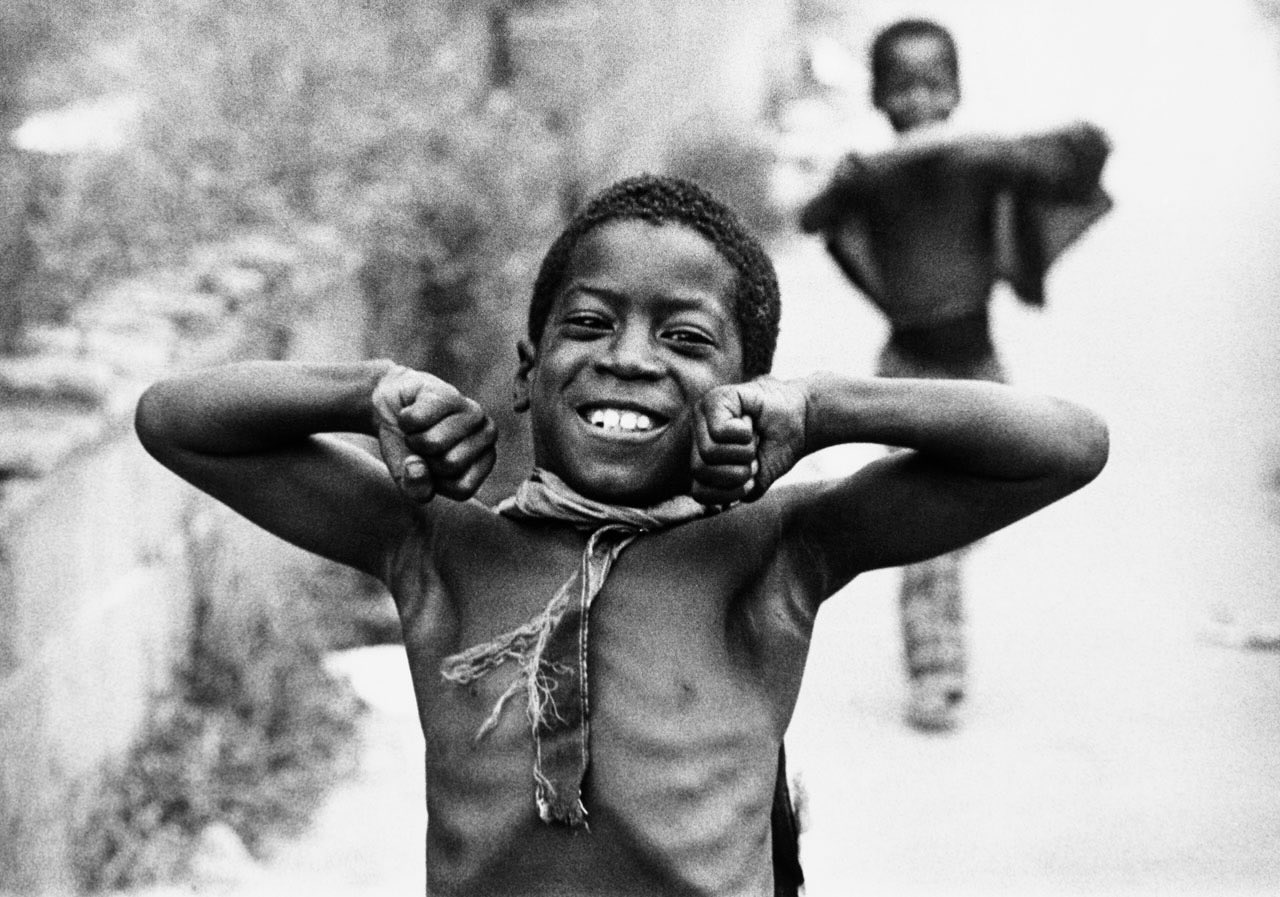 A black and white picture of a young Black boy on a residential street depicts the boy smiling at the photographer, fists on either side of his face. He is not wearing a shirt and is wearing twine and a furry pendant around his neck. Another boy is in the background.