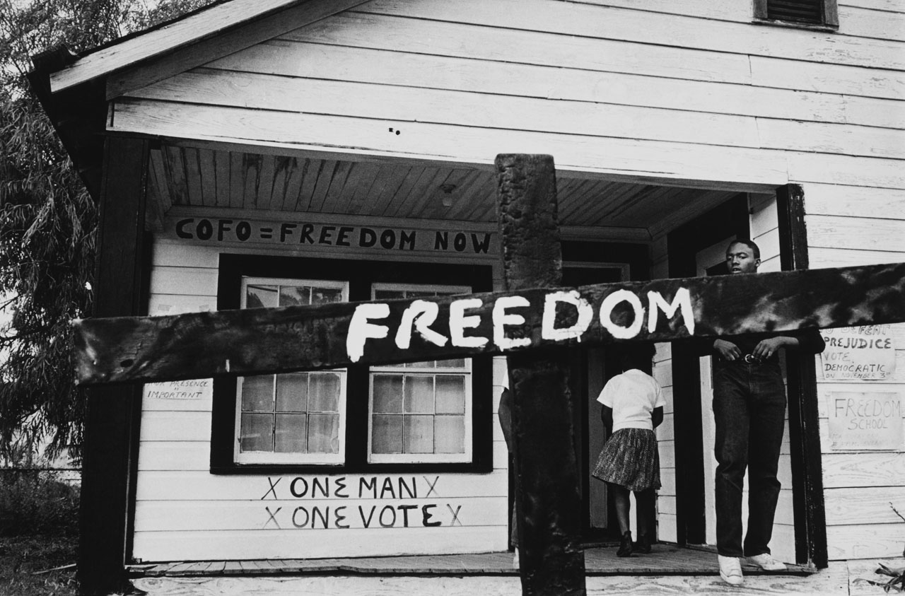 A black and white documentary-style photograph depicts a dark wooden fence with the word &lsquo;Freedom&rsquo; painted it on it. A young Black man stands behind the fence in the front entranceway of a house; behind him is another person stepping towards the door.