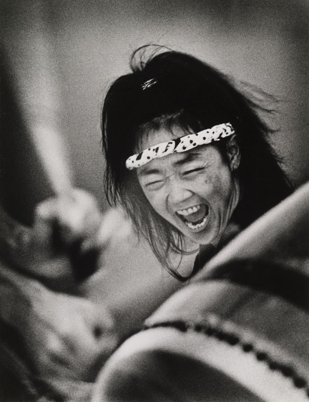 A black and white portrait of Linda Uyehara Hoffman, a woman with long black hair and a white patterned headband across her forehead, mid-shout or mid-laughter. In the foreground is a taiko drum.