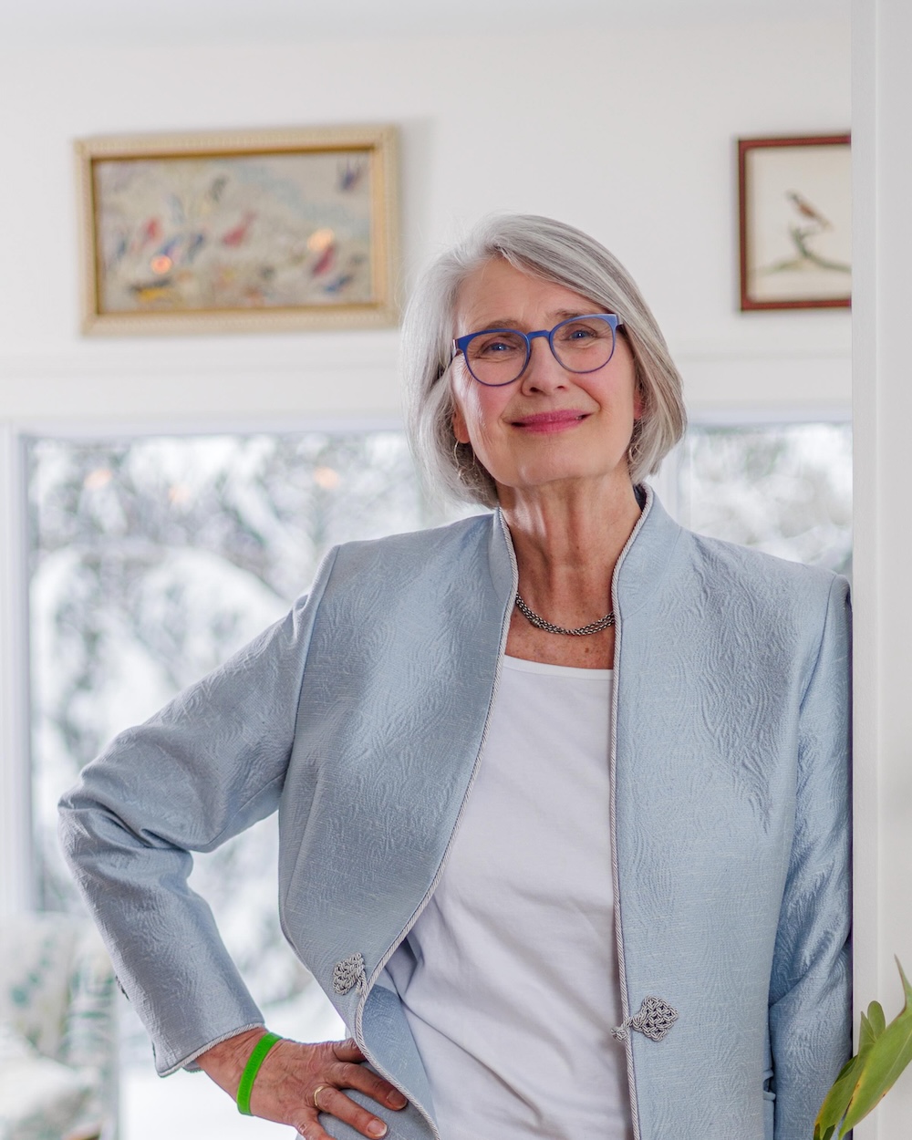 Louise Penny has chin-length grey hair and blue glasses. She is smiling at the camera and standing with a hand on her hip, wearing a light blue blazer over a white top. She is standing indoors in a brightly lit room with art in the background.