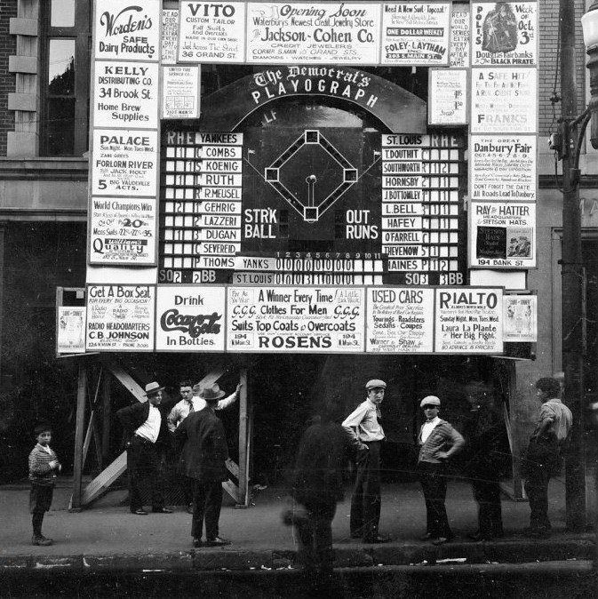 An old black and white photo of &lsquo;The Democrat&rsquo;s Playograph,&rsquo; consisting of a large raised board showing a baseball diamond with the inning-by-inning score underneath and players&rsquo;s names on either side. Advertisements frame the Playograph; one reads, &lsquo;Drink Coca-Cola in bottles.&rsquo; People in 1920s attire can be seen below the Playograph.