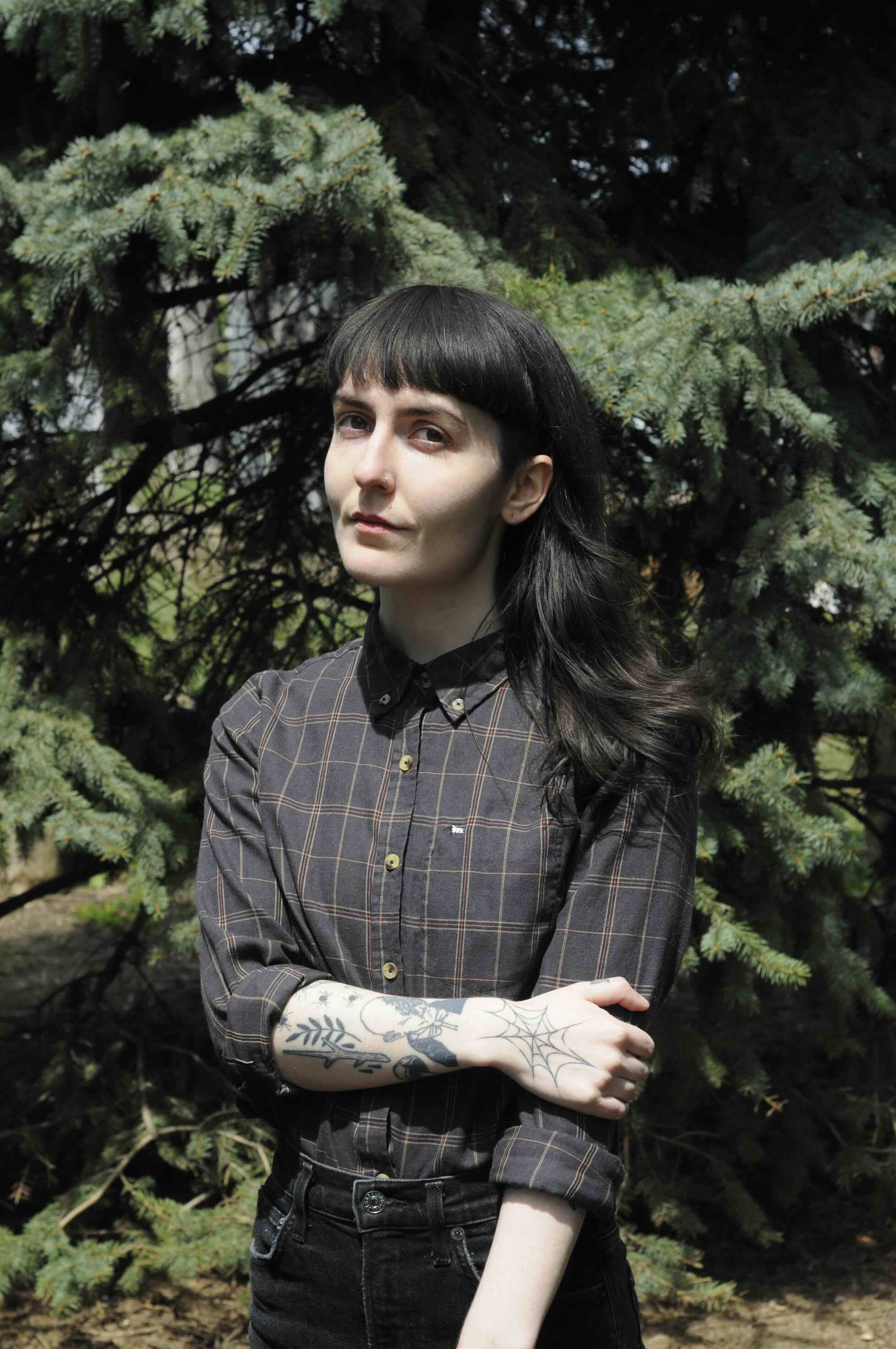 Sid Sharp has black dyed hair with bangs and a pale skin tone. They are wearing a black checked button-down shirt and black jeans. They&rsquo;re standing against a stand of pine trees.