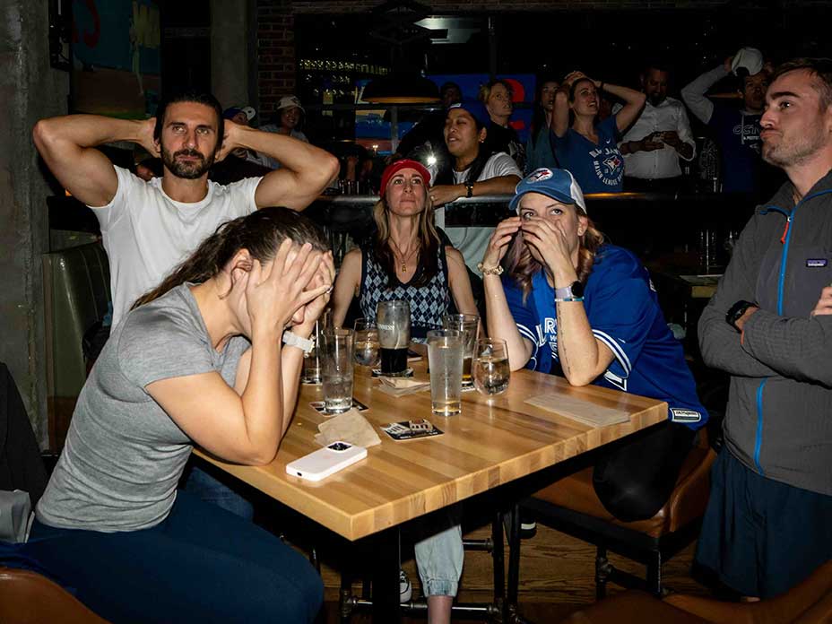 Five people are gathered around a small wooden table at a bar. A man stands to the left of the frame with his head in his hands, and the woman in front of him is seated with her face in her palms. Others at the table look on with apprehension.