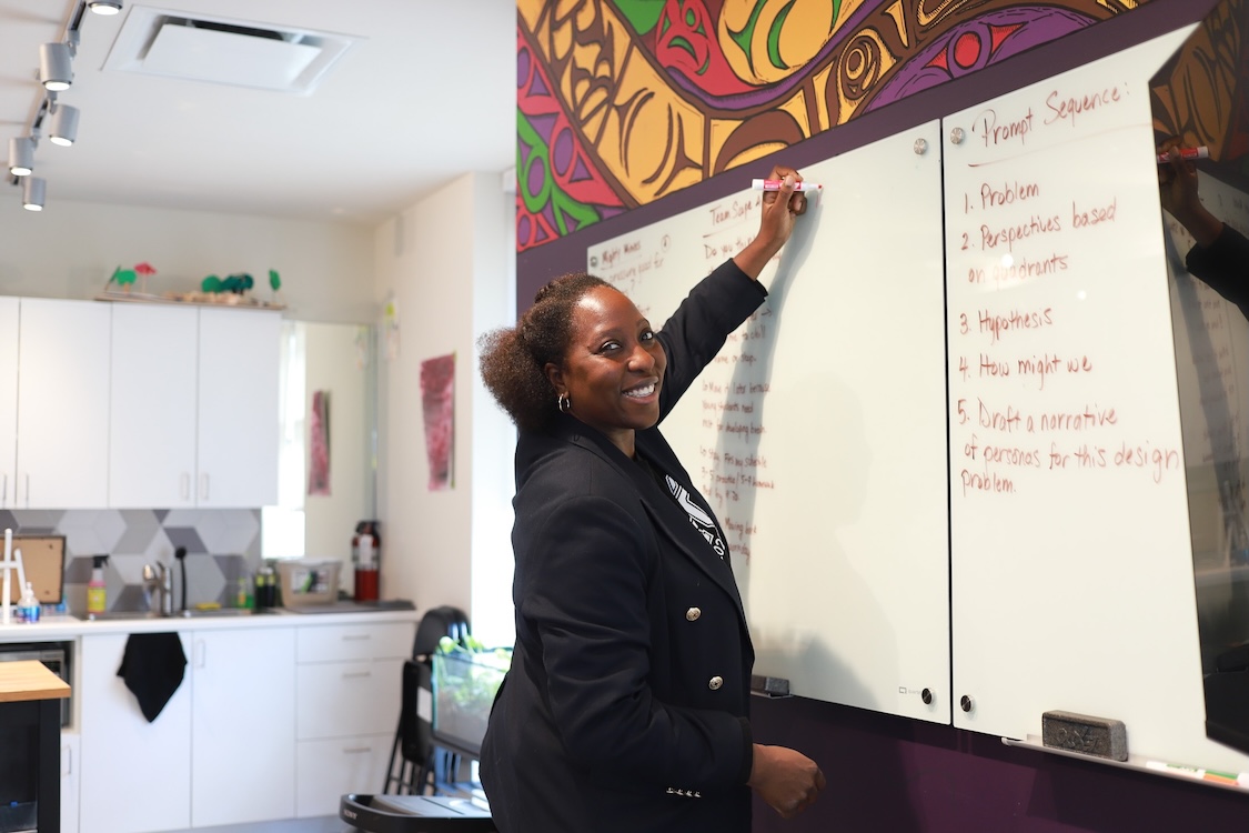 Anthonia Ogundele reaches up with her left hand holding a marker to write on a whiteboard installed against a wall featuring a colourful mural. She is a Black woman with short black wavy hair. She is wearing a dark blazer over a black T-shirt and smiling. Behind her to the left of the frame is a set of white cabinetry and a sink.