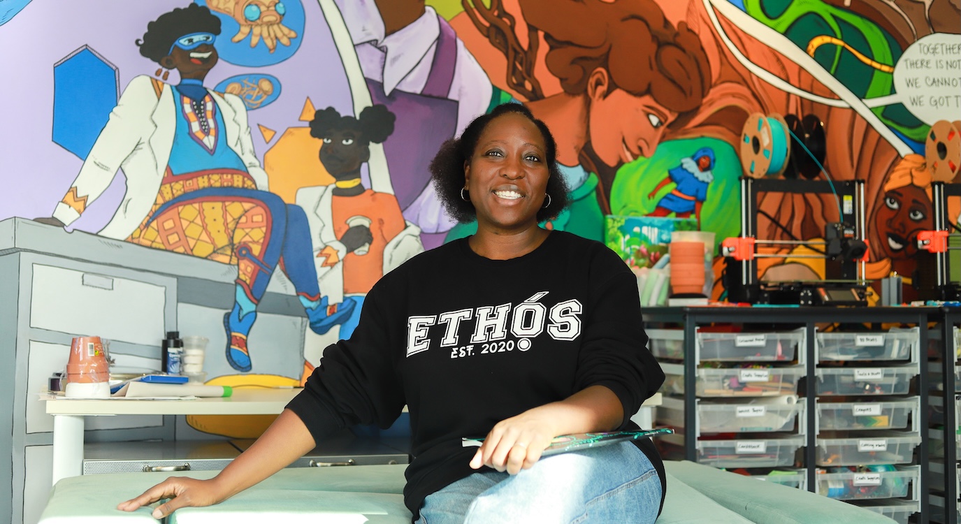 Anthonia Ogundele is seated on a green table in a laboratory with a colourful mural on the wall behind her. She is a Black woman with short dark wavy hair. She is wearing a black sweatshirt that says &ldquo;Ethos&rdquo; in bold white text and light blue jeans.
