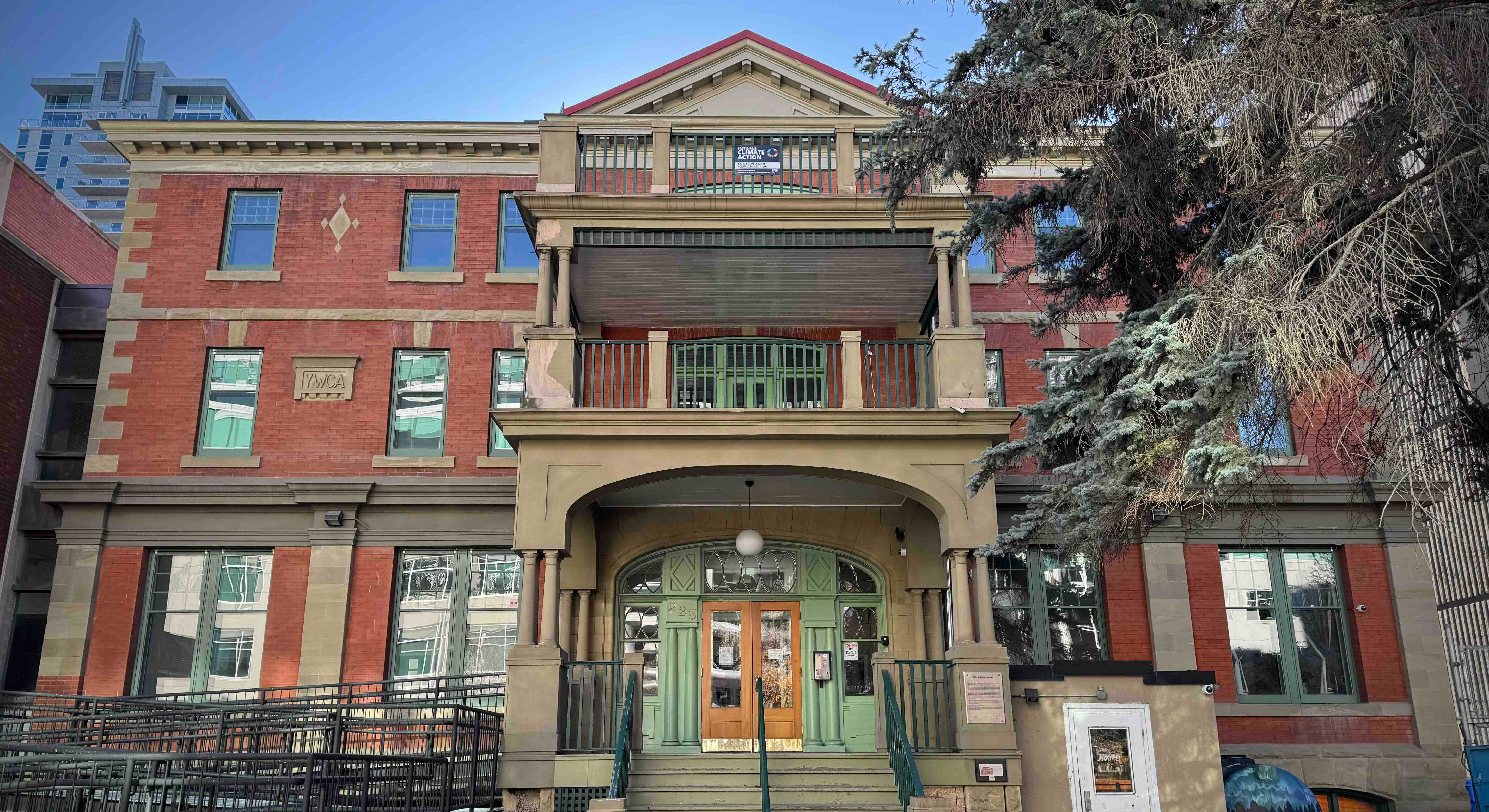 A red brick Georgian Revival building stands on a Calgary street on a clear day. To the right of the frame are the branches of a coniferous tree.