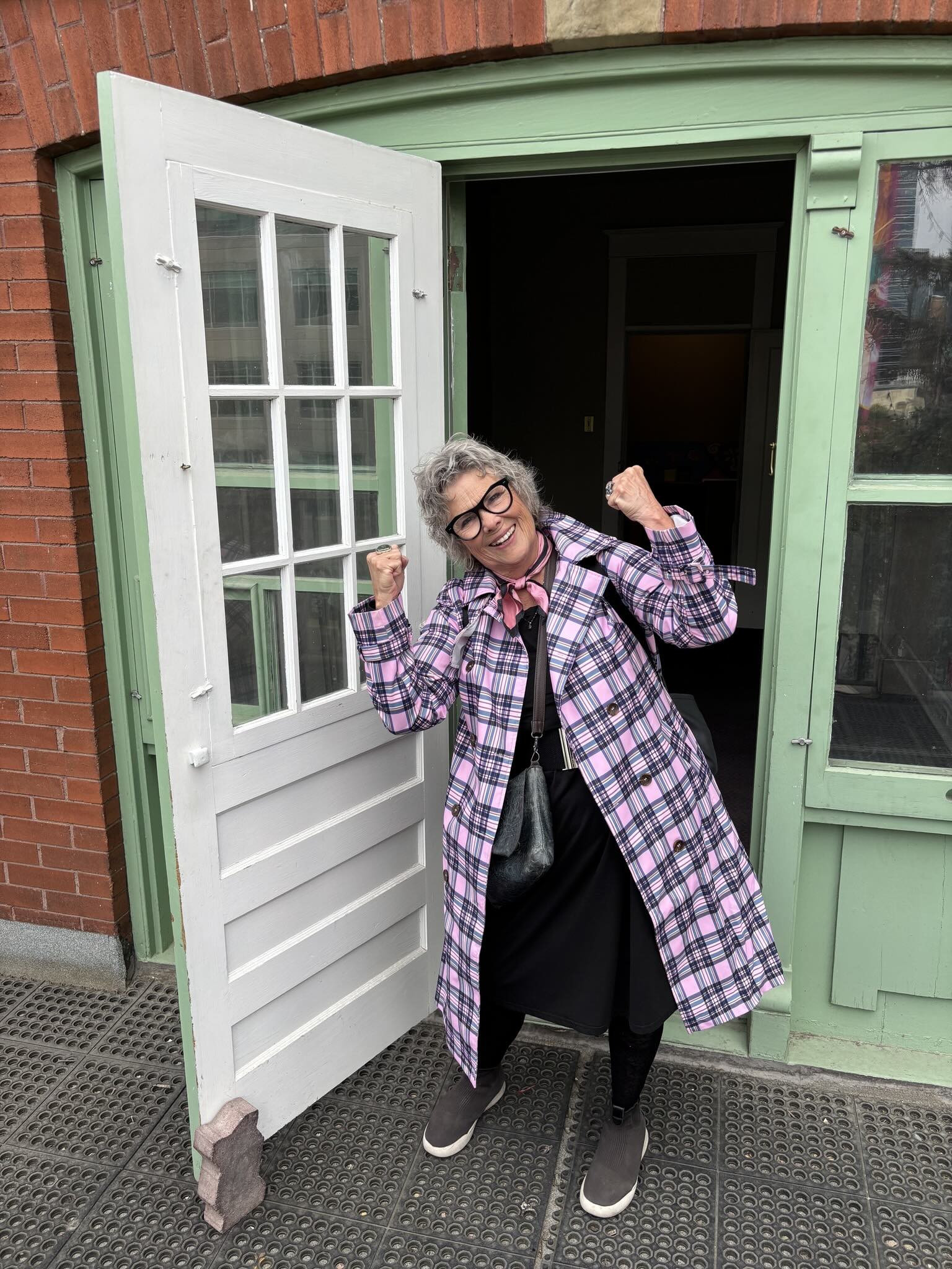 Sharon Stevens has short wavy grey hair, thick framed glasses and a light skin tone. She is wearing a bright pink plaid jacket over a black dress and standing with her fists up in the green and white doorway of a brick building.