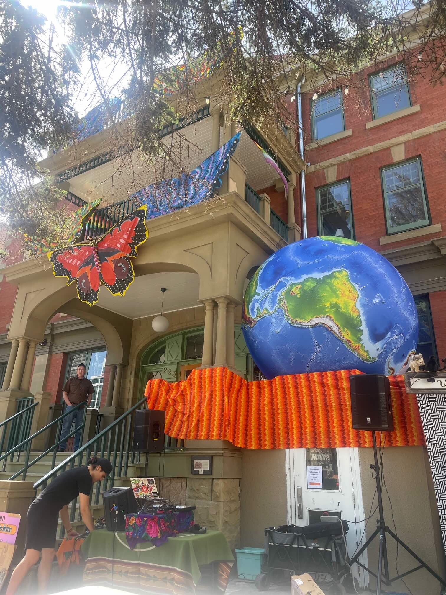 A large butterfly and planet earth installation decorate the Georgian Revival façade of an historic building while one person stands on the porch and another person sets up items on a table at the bottom of the front steps. 