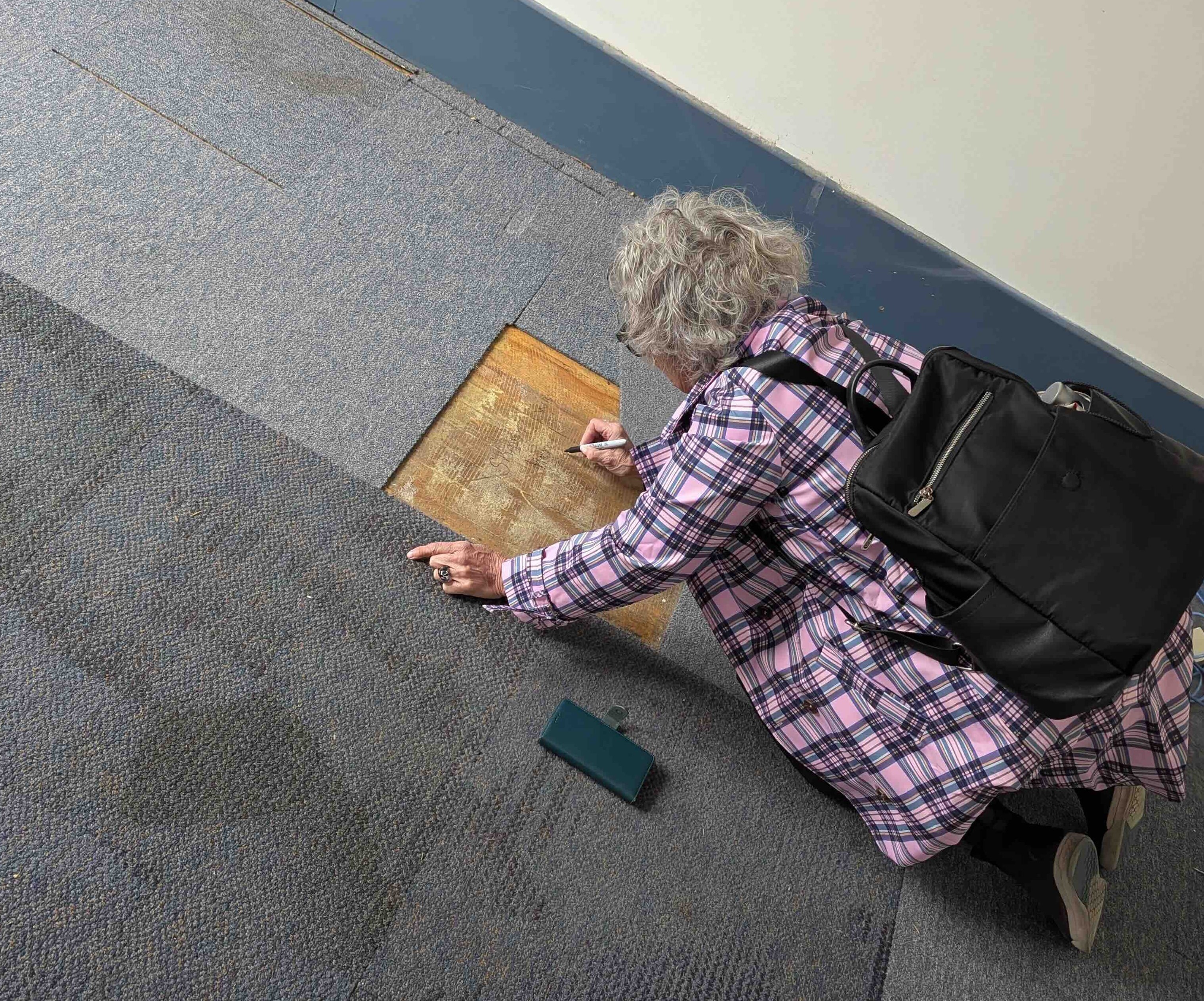 Sharon Stevens has short wavy grey hair. She is wearing a long pink plaid jacket and a black backpack. Her back is turned to the camera and she kneels on a carpeted floor to sign her name on a piece of wood under the carpet.
