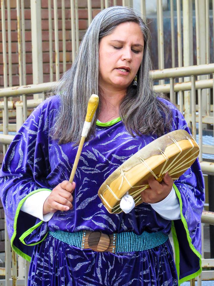 Michelle Robinson has long dark greying hair and a medium skin tone. She is wearing a purple top, a turquoise beaded belt and she is playing a small circular drum.