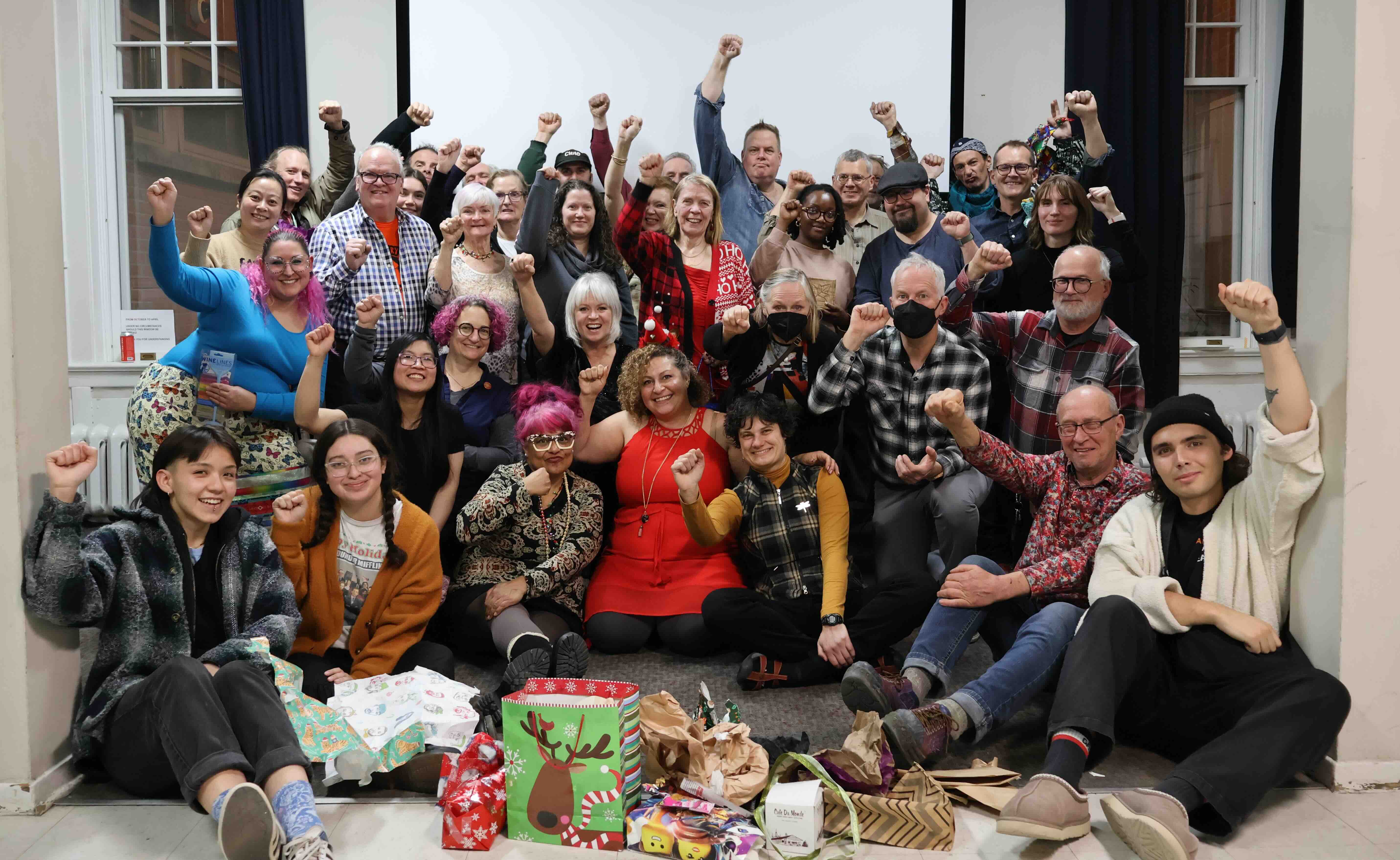 A large crowd of people is gathered in an indoor party room. Some are seated on the floor and others are standing. They are holding a fist up in solidarity, smiling.