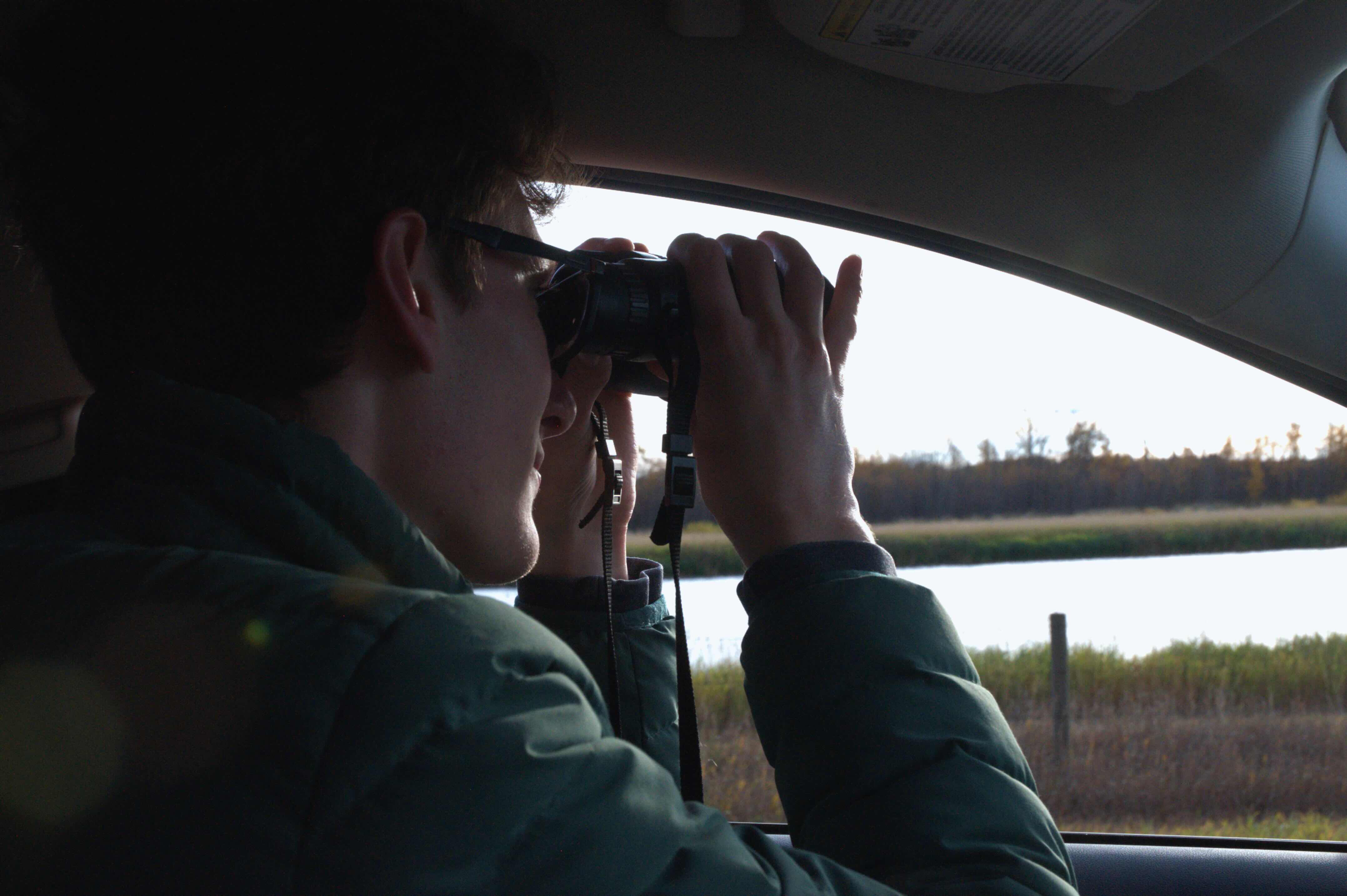 David Grinevitch is seated in the driver’s seat of a car and looking through binoculars at a body of water outside. He is wearing a green puffer jacket and has short brown wavy hair.