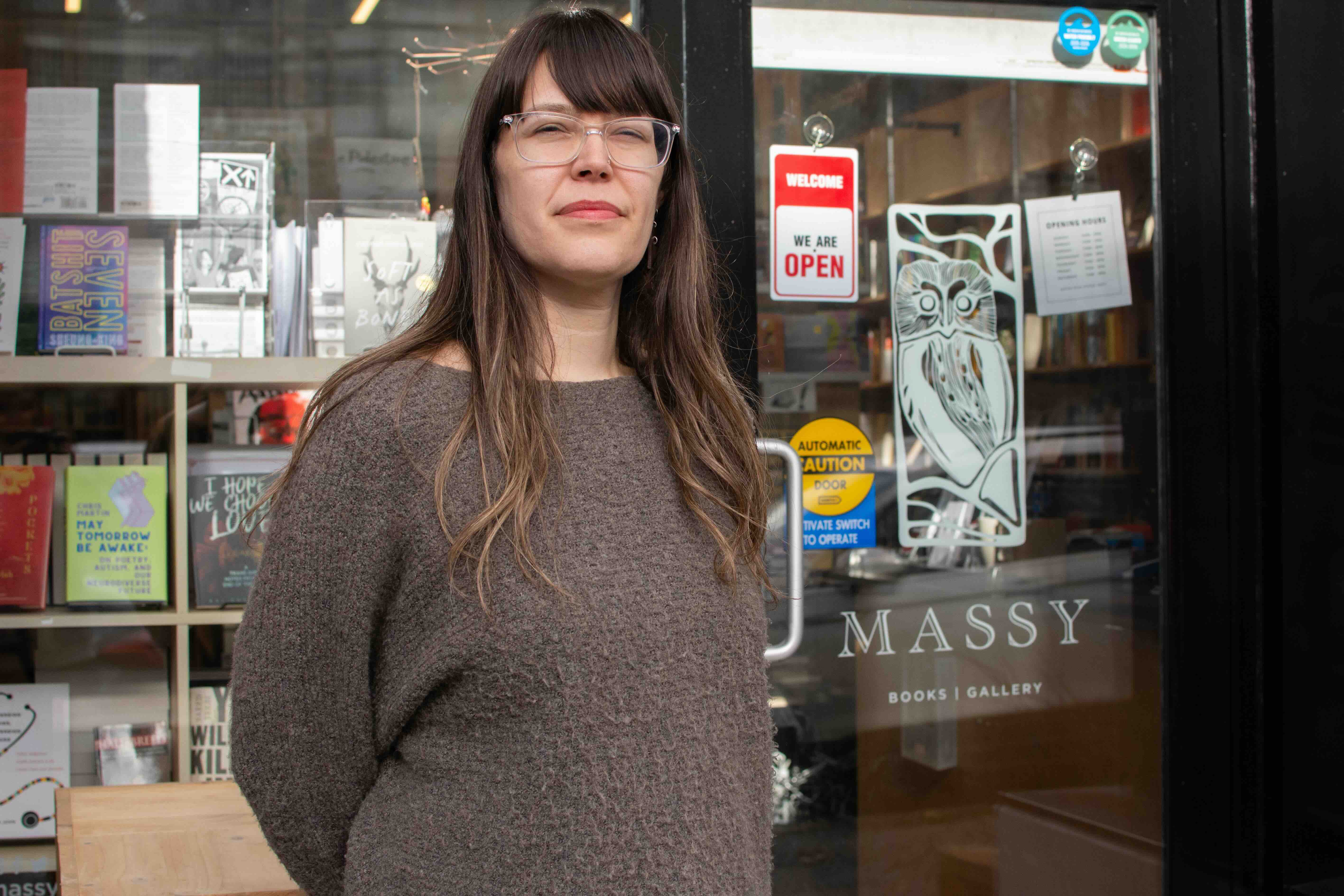 Patricia Massy has long wavy brown hair. She is wearing glasses and a brown sweater, and she is standing outside the front door of her bookstore which has a piece of white owl artwork etched against the glass below text that reads &ldquo;Massy Books: Books | Gallery.&rdquo;