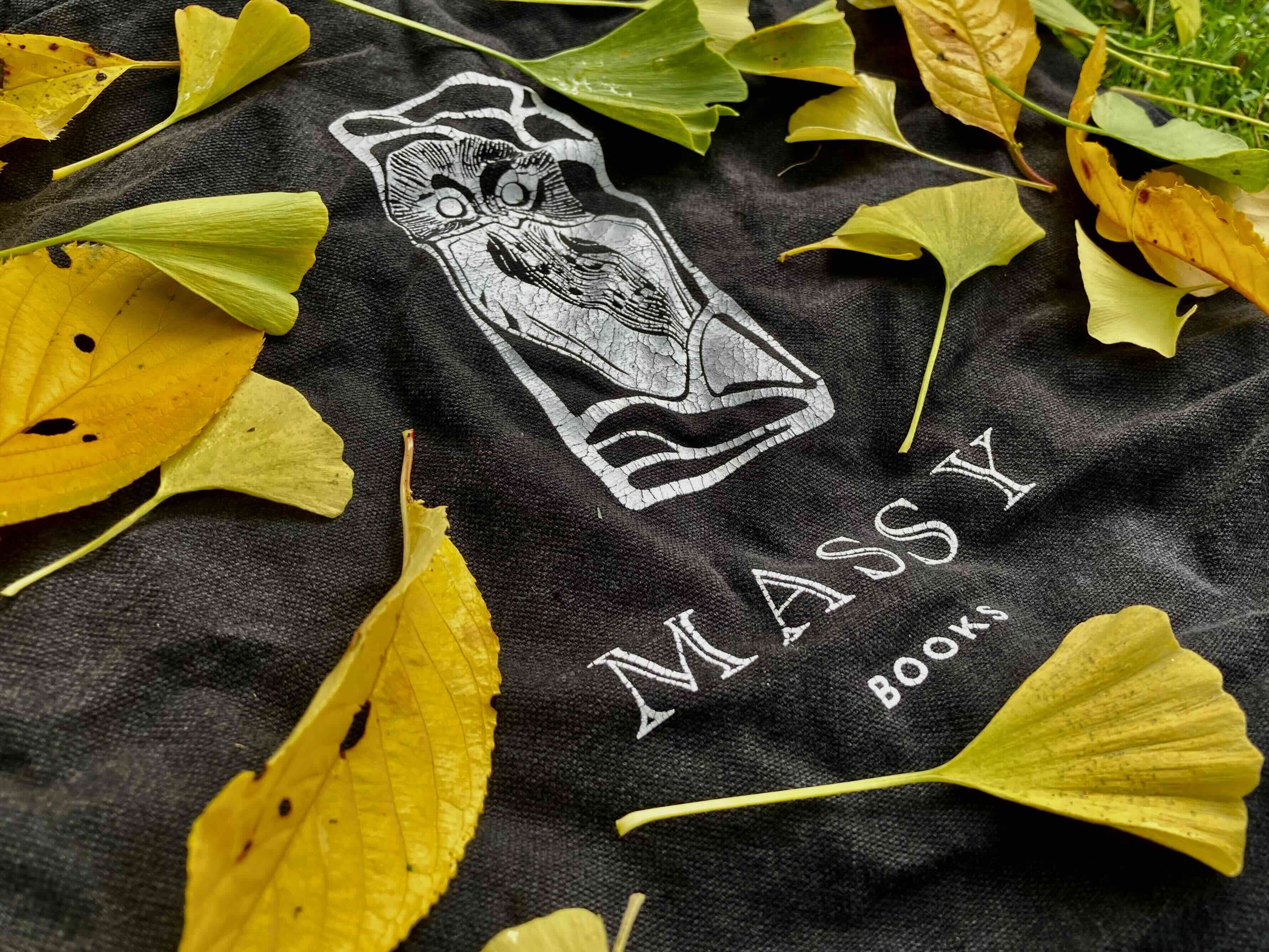 A black tote bag with a white graphic of an owl and the words &ldquo;Massy Books&rdquo; is arranged on grass under fall leaves.