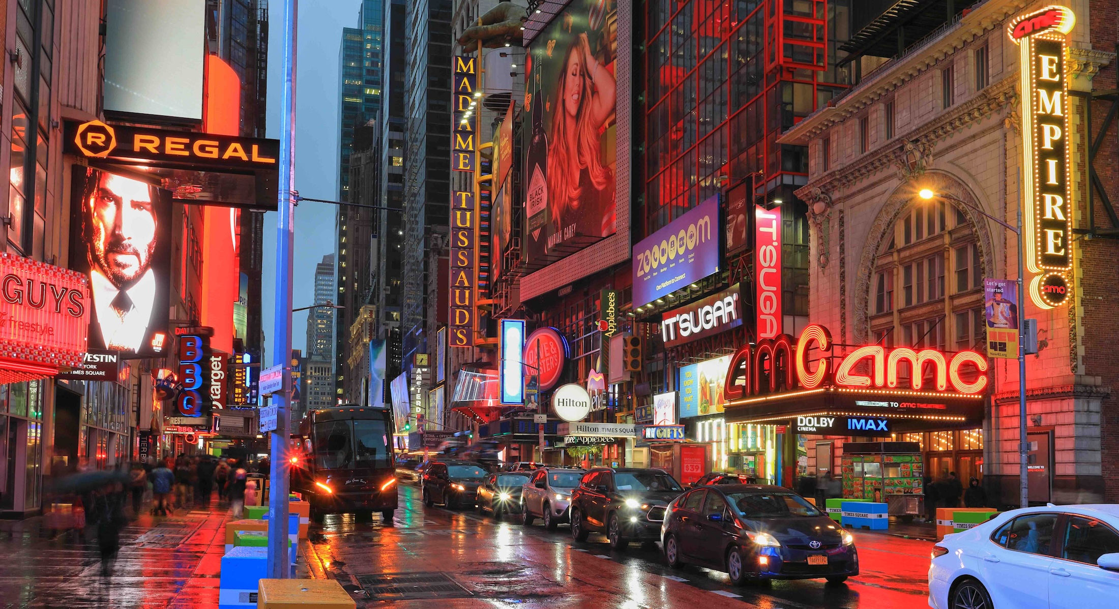 A photograph of a busy downtown street in New York City&rsquo;s theatre district features cars and pedestrians travelling at dusk on a rainy night. Bright lights from theatre marquees line the street.