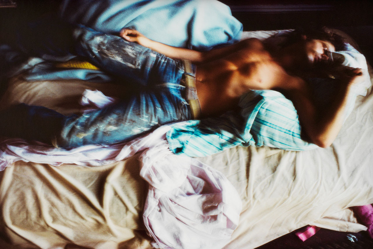 A shirtless man in blue jeans lies across a bed with rumpled pink, beige and light blue sheets.