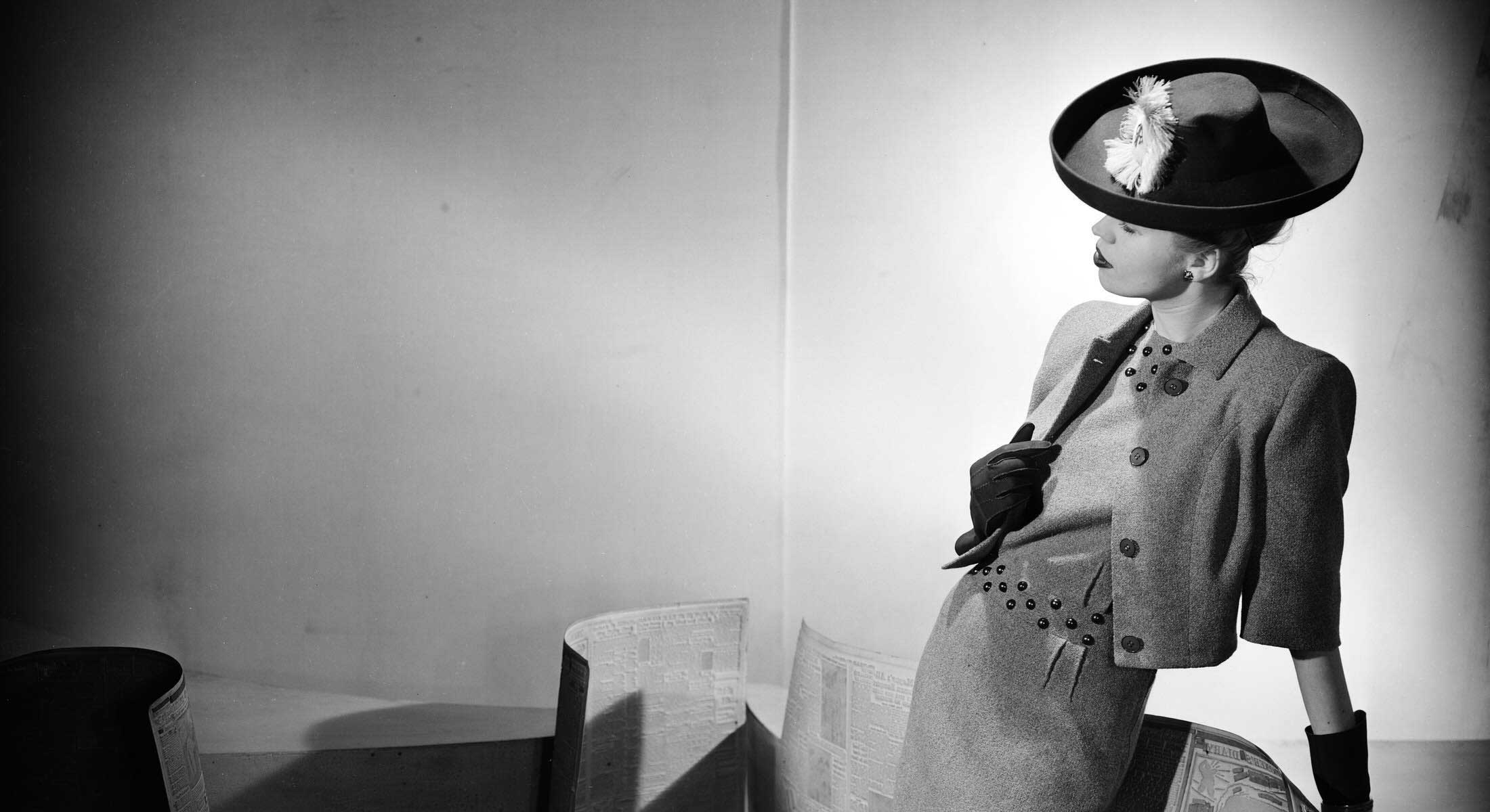 A woman poses by leaning against the back of a sofa. She is wearing a dark feathered hat and a short tweed jacket over a matching dress in a 1940s style. She is in an indoor space and low tables and a black wall are to the left of the frame.