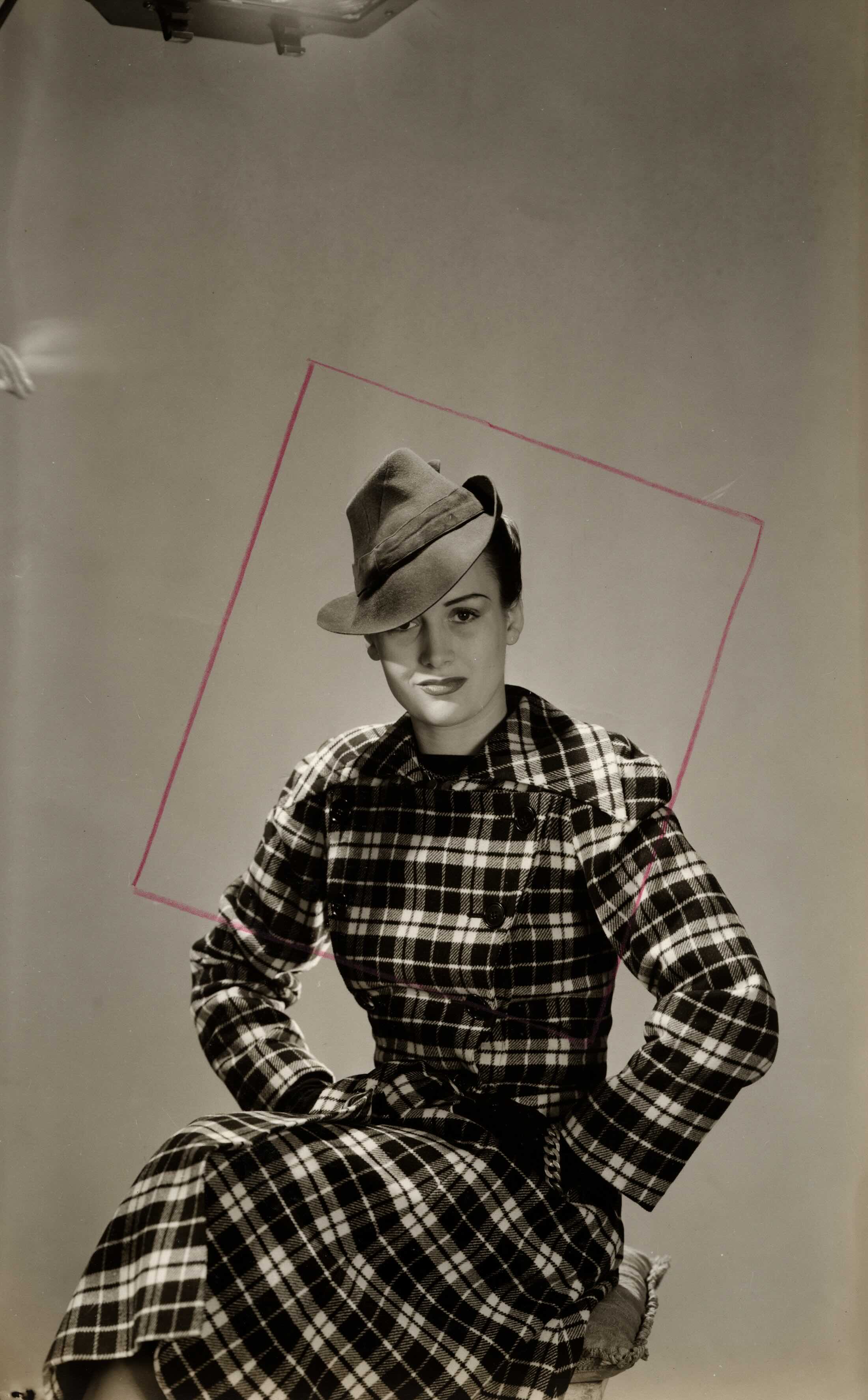 A woman in a plaid trenchcoat is seated against a studio background wearing a 1940s style fedora.