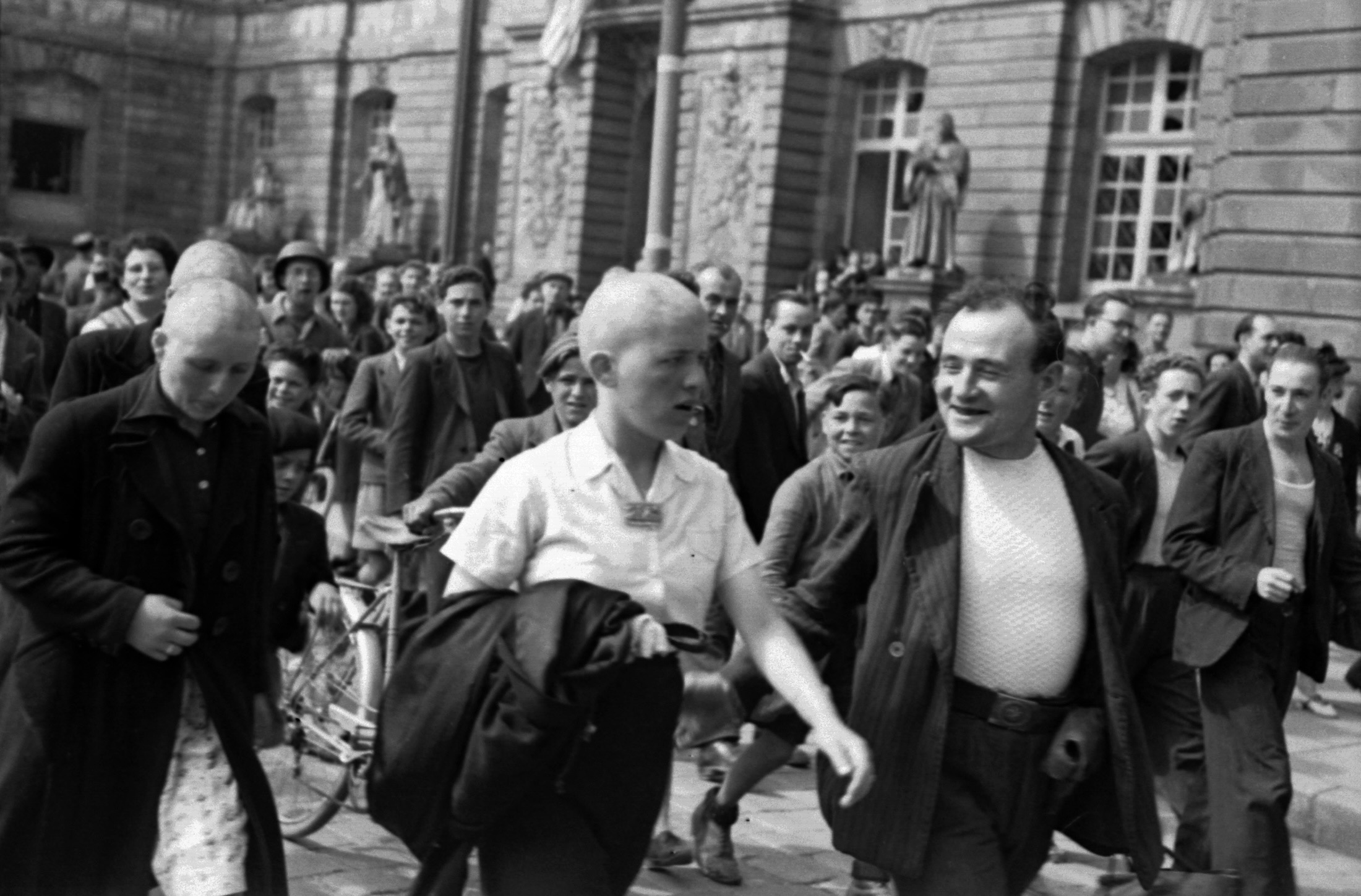 A woman with a shaved head and a short-sleeved white shirt is walking down a crowded street full of people. A man to her right is trying to speak to her. Behind her are other women with shaved heads.