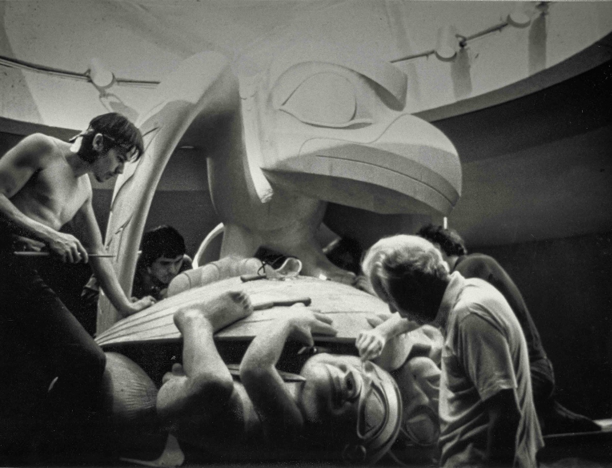A black and white photograph depicts several men working together on a piece of Haida artwork.