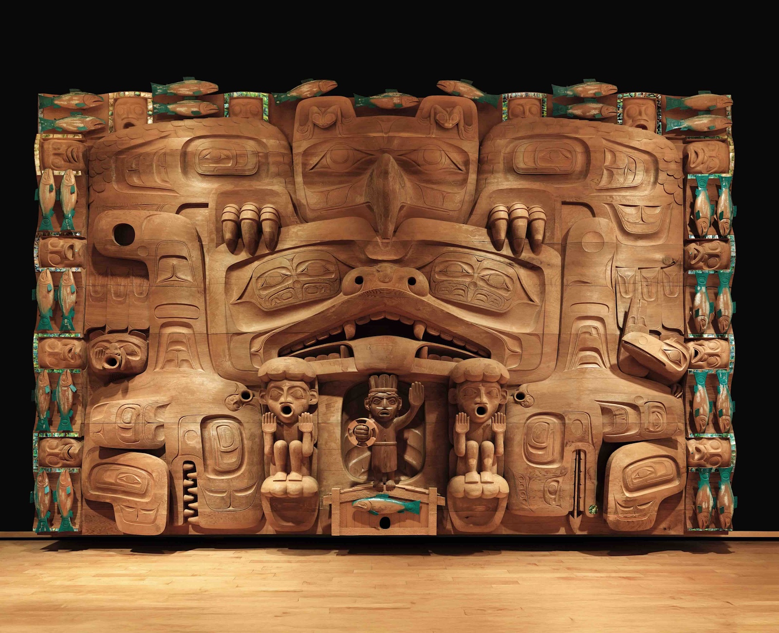 A large-format Haida wood carving against a black background on a wood floor with turquoise details.