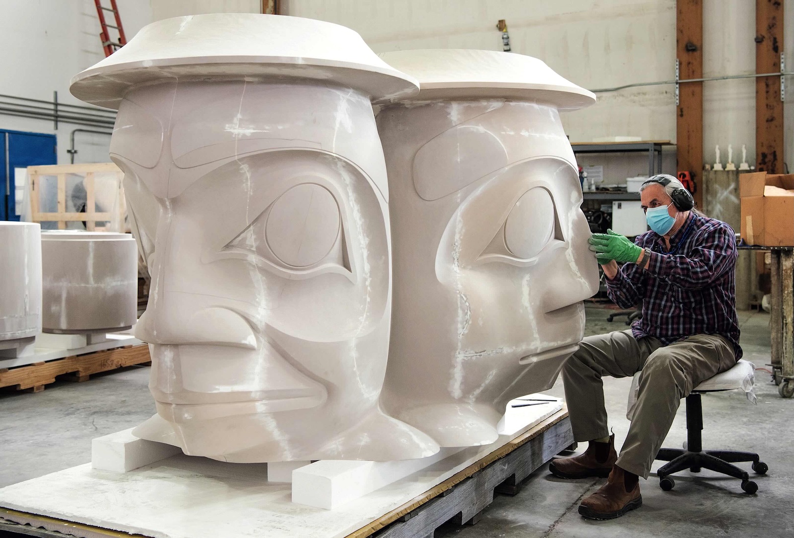 James Hart is seated to the left of the frame in a blue and red plaid shirt and olive-coloured trousers. He is wearing a blue medical mask, black ear protectors and green gloves while he works on a large-format carving of three faces, which are of a solid shiny white material. Hart is working in a large indoor studio space. 