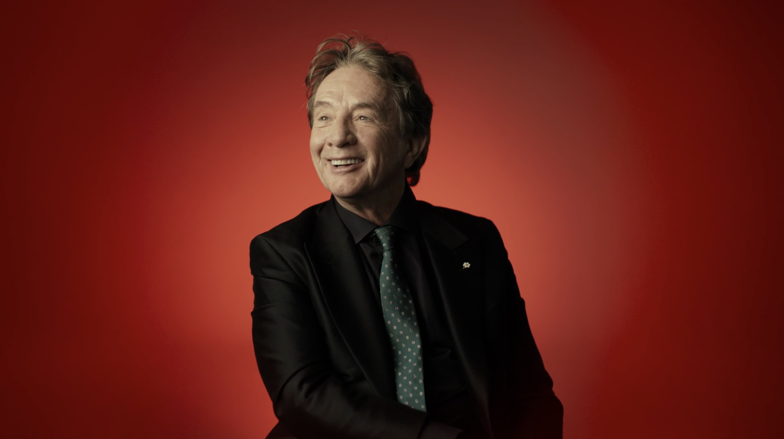 Martin Short is wearing a black blazer over a dark shirt and green tie. He is looking to the left of the frame, smiling and seated against a red background.