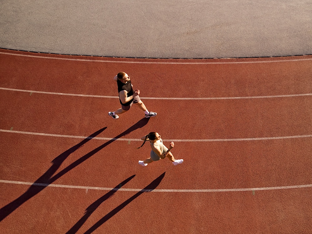 A bird&rsquo;s-eye view of two people jogging on a brown track with white stripes. They both have a pale skin tone and blond hair.