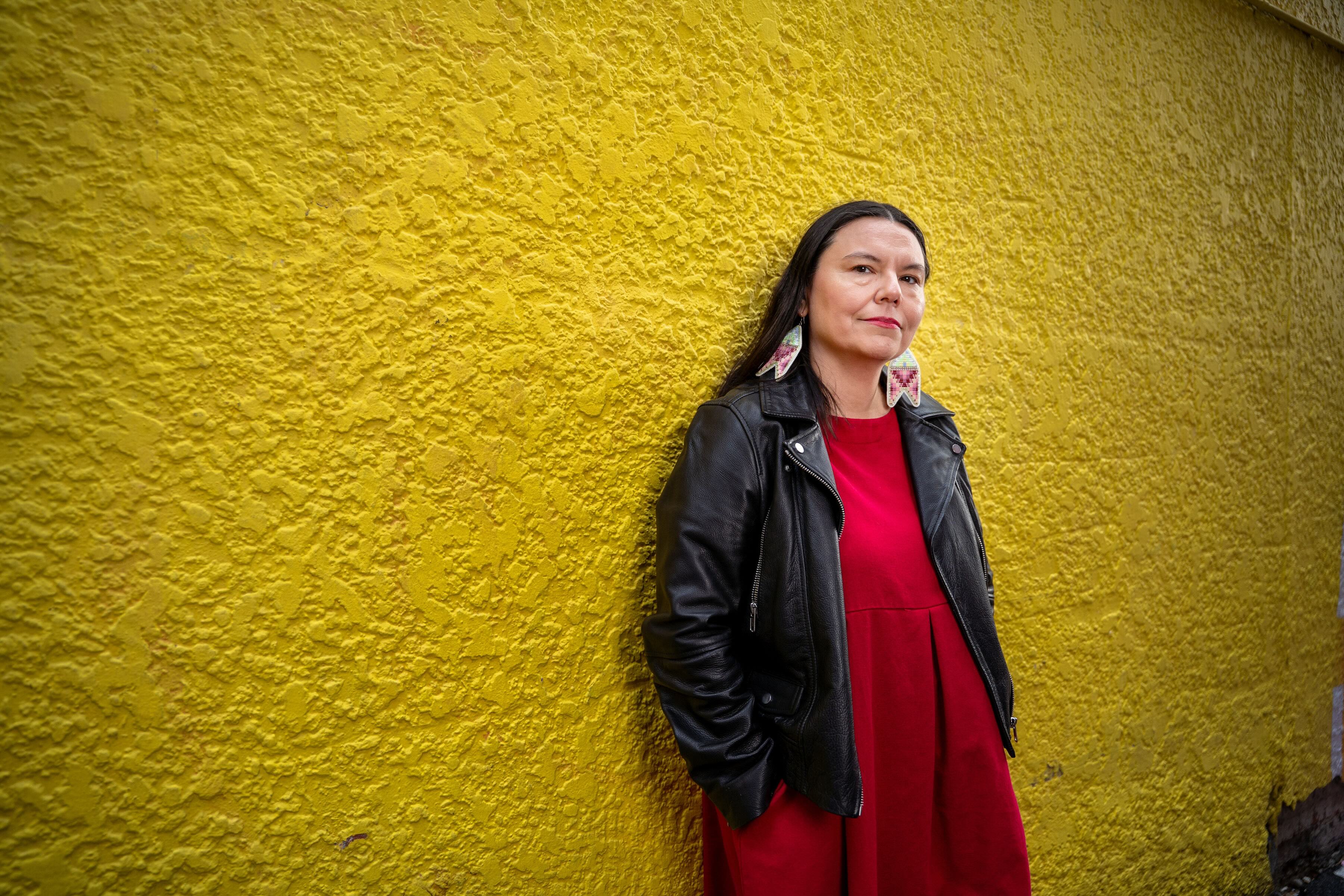 Eden Fineday stands against a textured yellow wall. She has long dark hair and is wearing colourful beaded earrings, a black leather jacket and a red dress.
