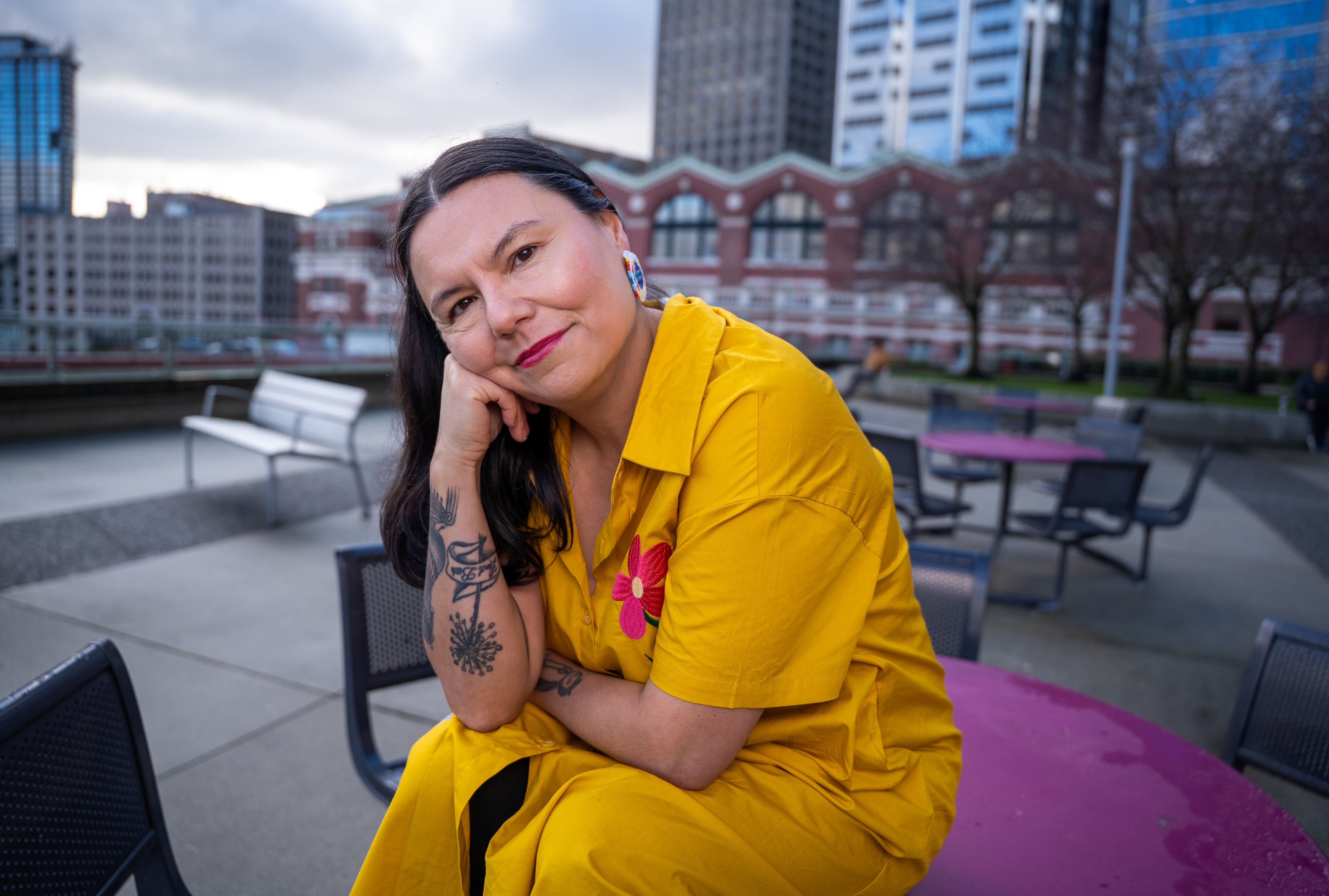 Eden Fineday is sitting on a magenta table in an outdoor plaza on a grey day. She is posing by leaning her cheek on her hand, and she is smiling. She is wearing a yellow dress with floral embroidery and round beaded earrings. She has long dark hair pulled back.