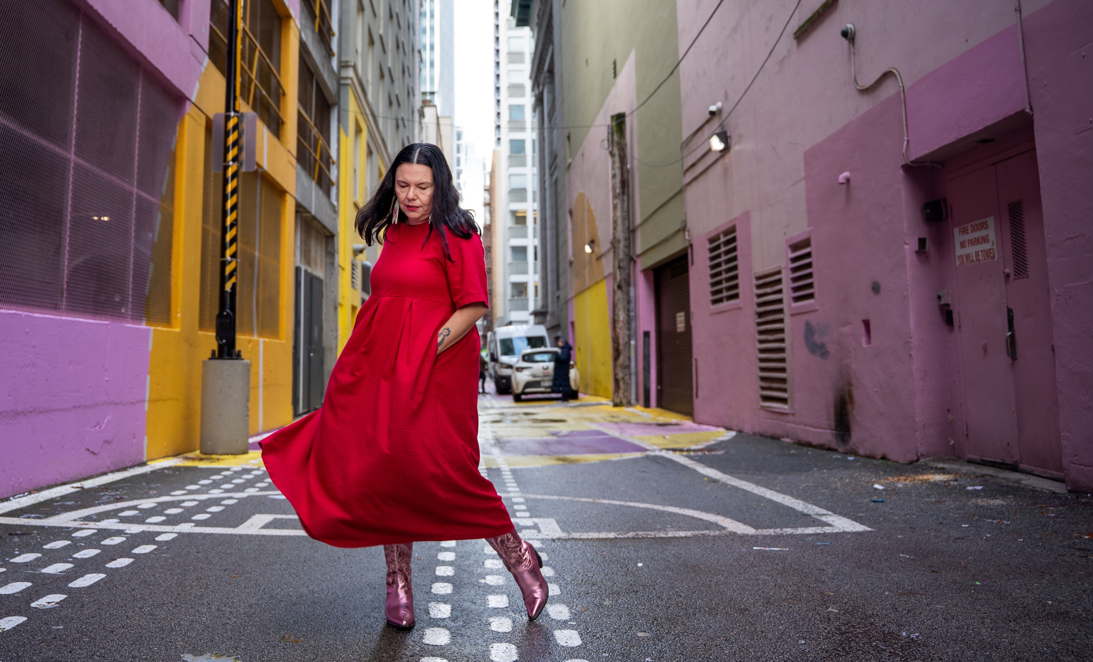 Eden Fineday has long dark hair. She is wearing a red dress and pink cowboy boots. She is standing in the middle of an alley with its walls painted pink. She is looking down, mid-step, with her hands in her pockets.