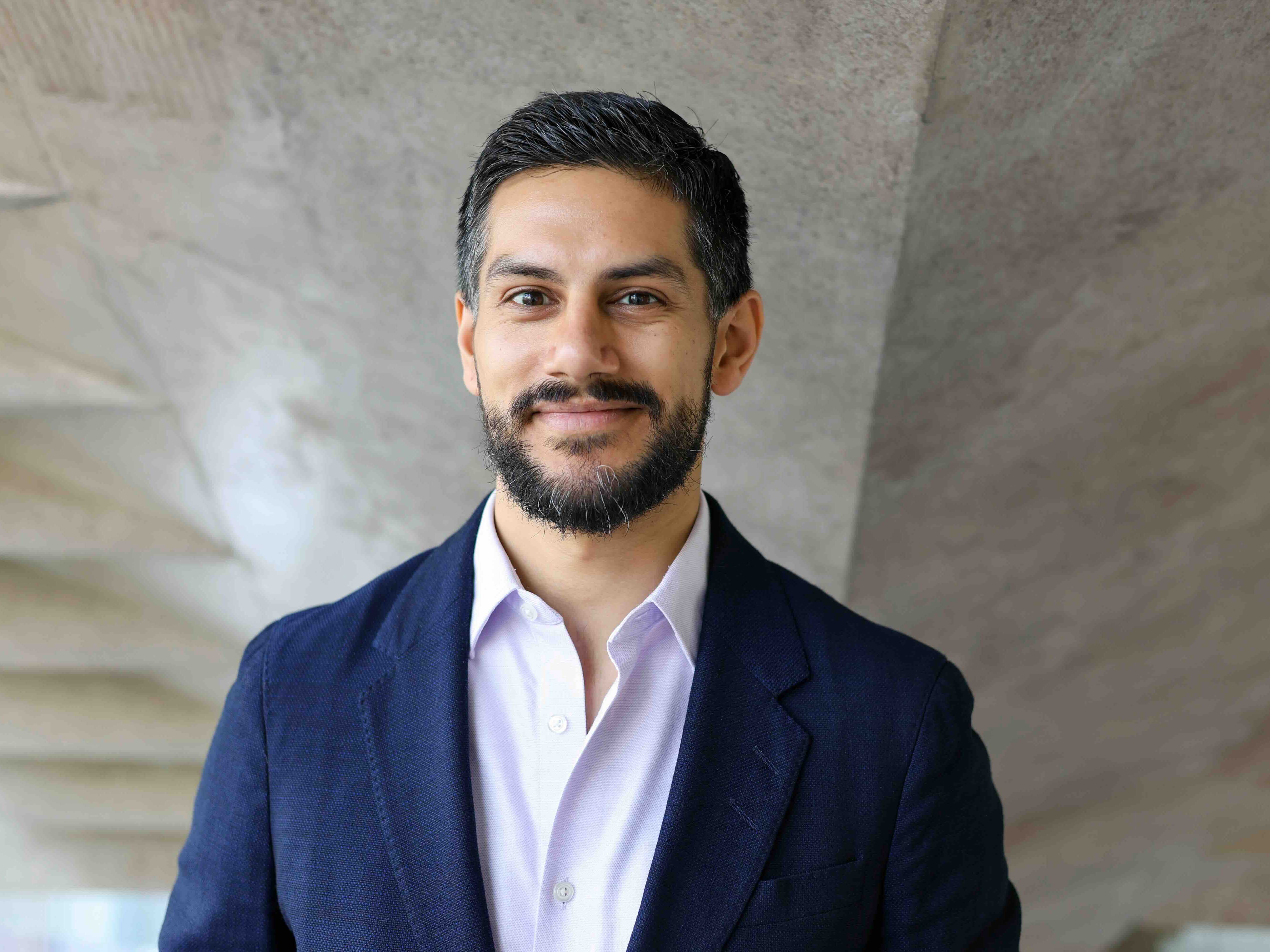 Liam Midzain-Gobin has short dark hair, a beard and a medium skin tone. He is wearing a navy suit and a lavender shirt. He is photographed against a marbled grey studio background.