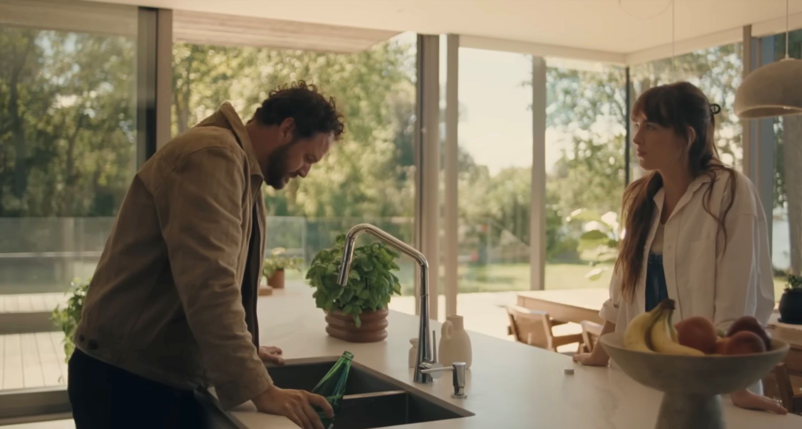A man stands to the left of the frame, looking down while leaning over a kitchen sink with white kitchen counters. Across from him to the right of the frame is a woman standing, looking at him. They both have brown hair and are wearing casual clothes.