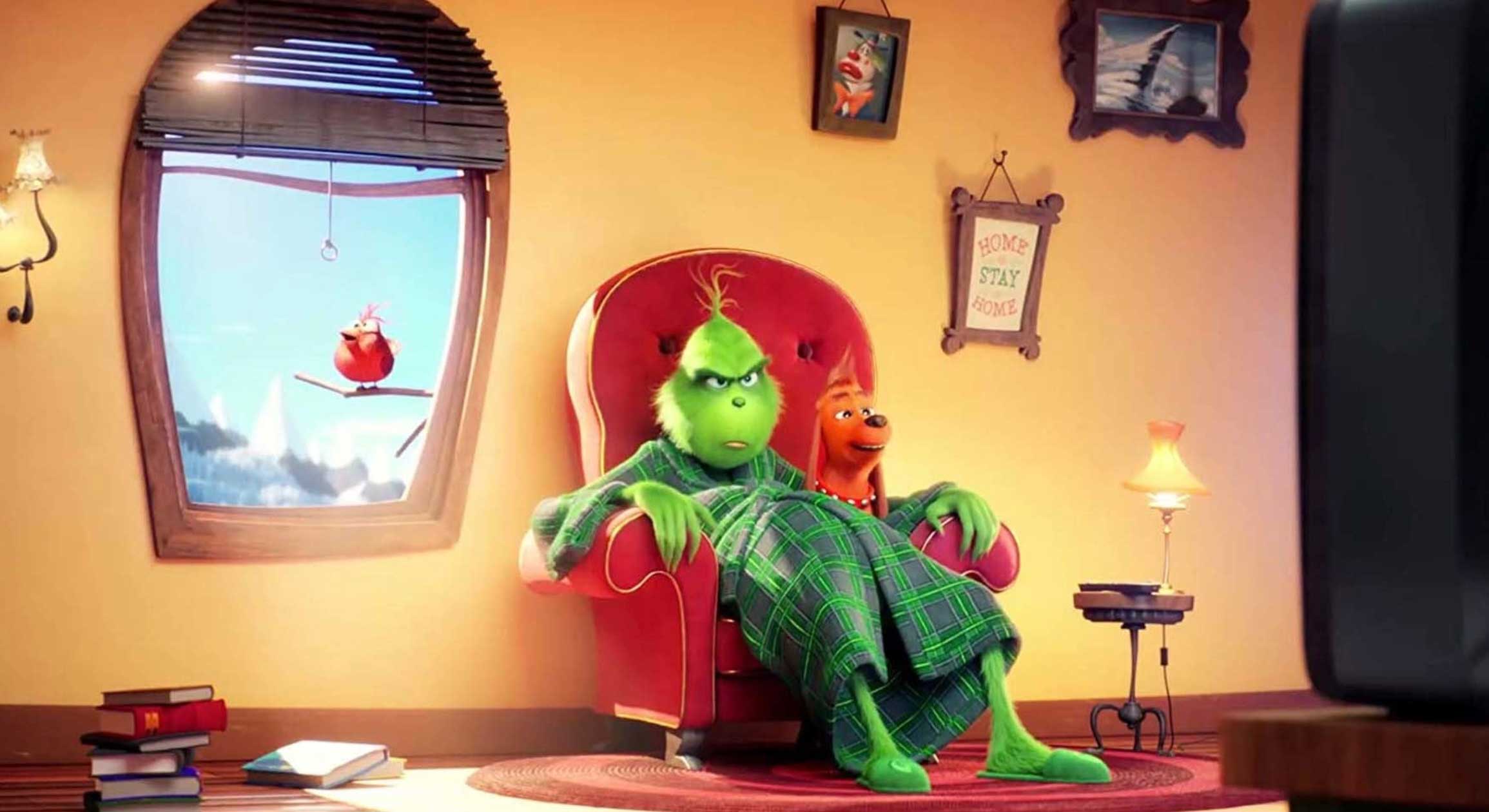 A digital still from 2018&rsquo;s &lsquo;The Grinch&rsquo; depicts a furry green creature sitting in a red armchair with a smiling dog at his side. He is wearing a green plaid bathrobe and a framed embroidery that says &ldquo;Home stay home&rdquo; is to his right.