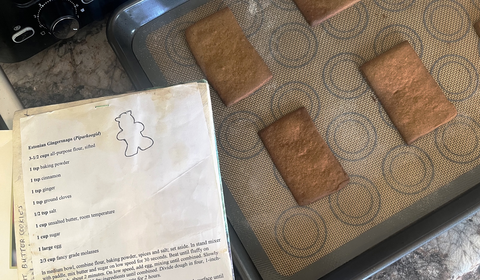 The cookie recipe in a little handwritten cookbook, beside a baking tray that has baked rectangular cookie pieces.