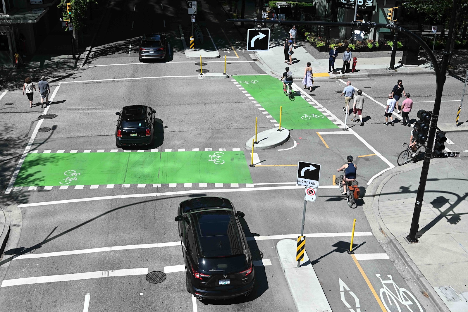An aerial view of a Vancouver street features two black vehicles driving through an intersection while pedestrians and cyclists cross in marked pathways on the right.