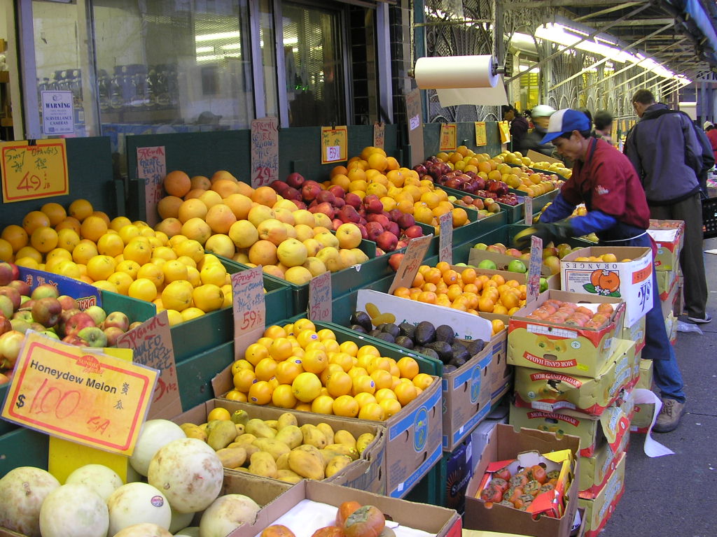 A man in a blue ball cap and red shirt stands to the right of the frame, loading fruits from cardboard boxes into the outdoor stand of a produce market. Behind him are customers shopping.
 